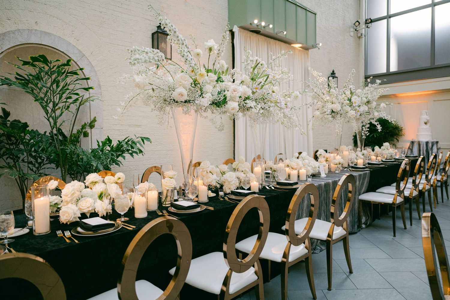 Modern wedding reception setup with white floral centerpieces and gold chairs in an elegant white space.