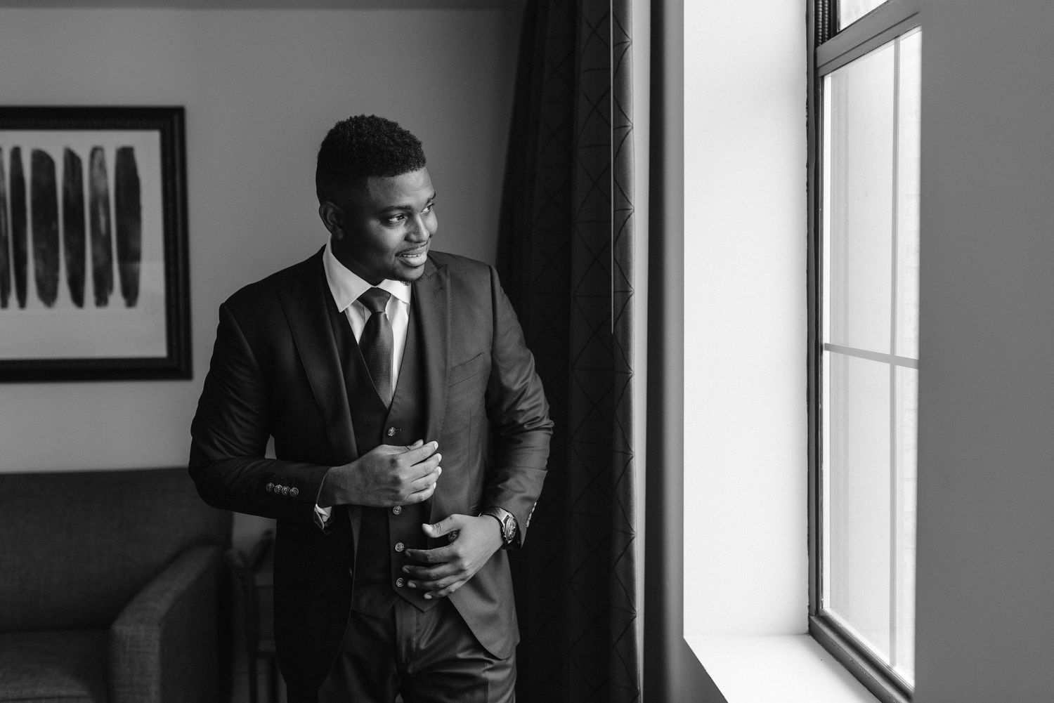 A groom in a dark suit stands thoughtfully by a window in black and white.