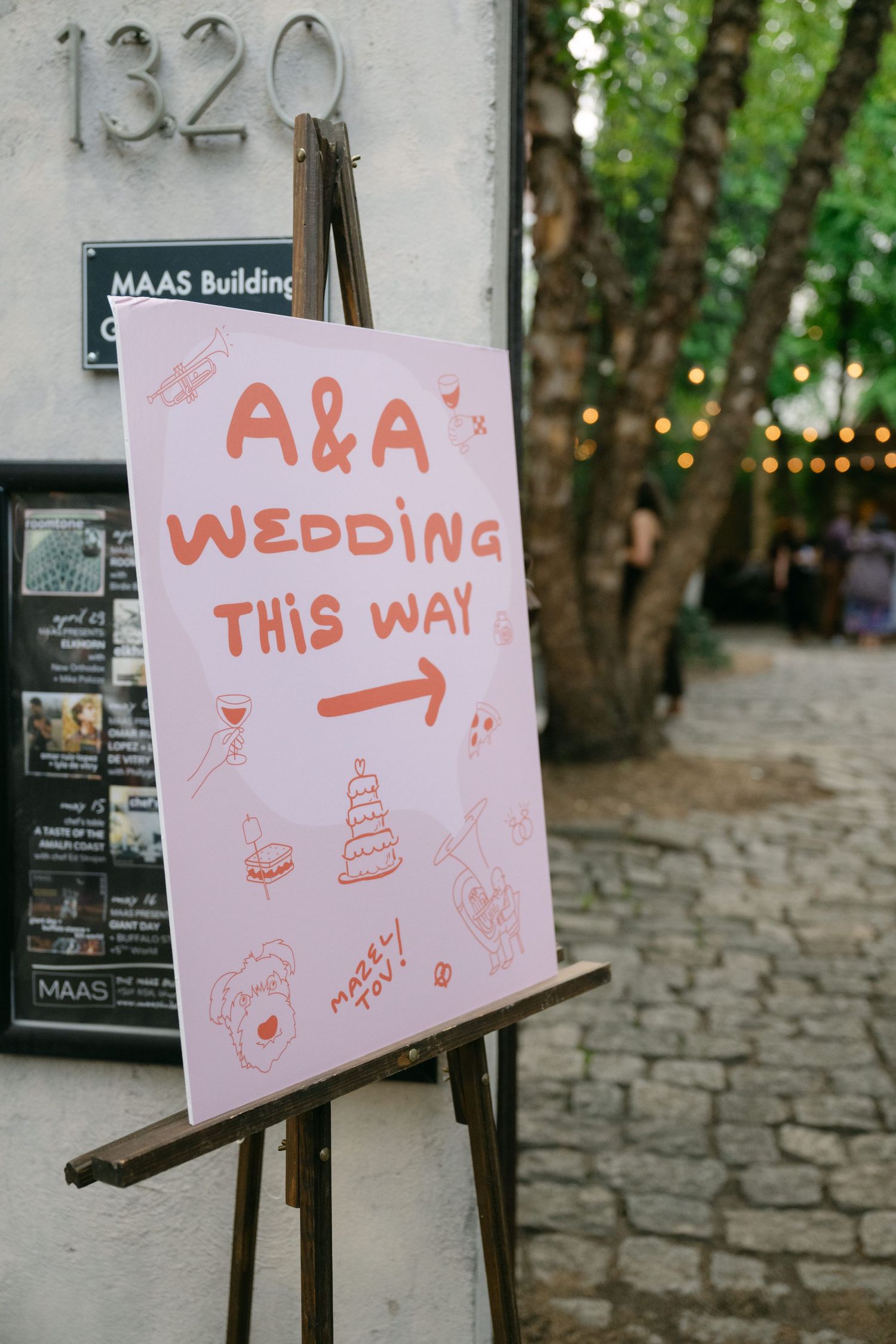 Pink handwritten wedding direction sign outside the MAAS Building with decorative doodles.