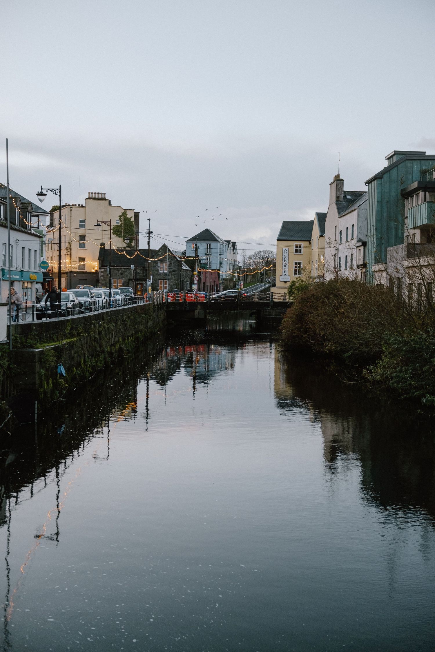 A tranquil canal reflects colorful buildings and bridges in an Irish town under an overcast sky.