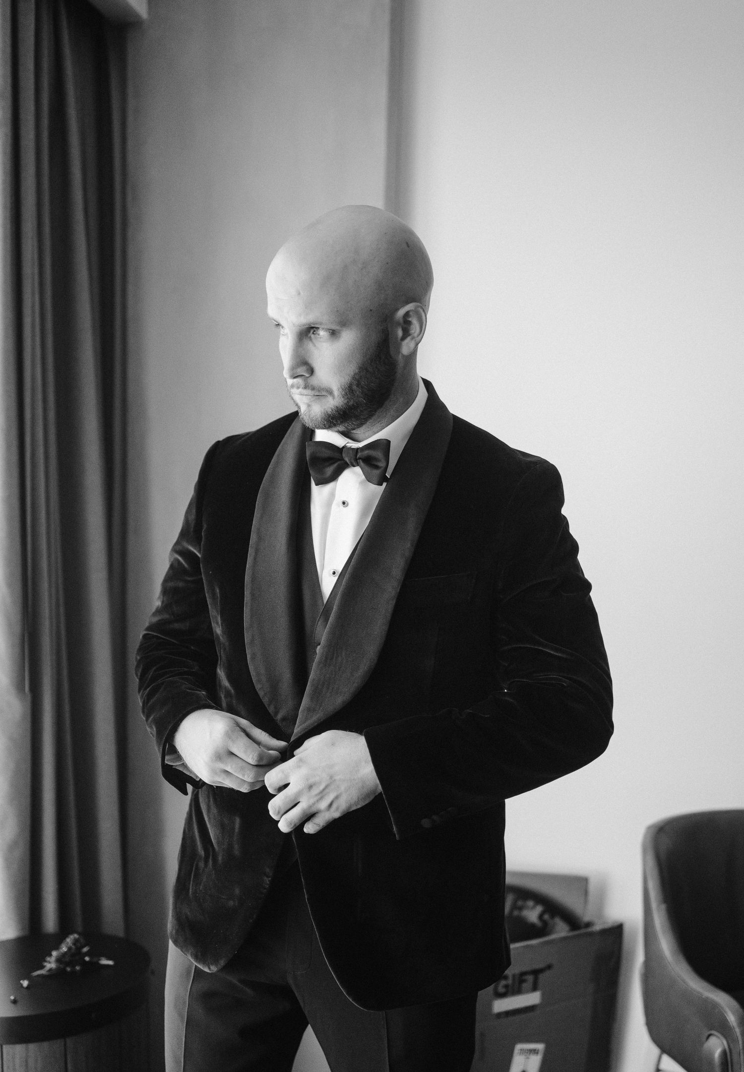A black and white portrait of a groom adjusting his bowtie in a velvet tuxedo.