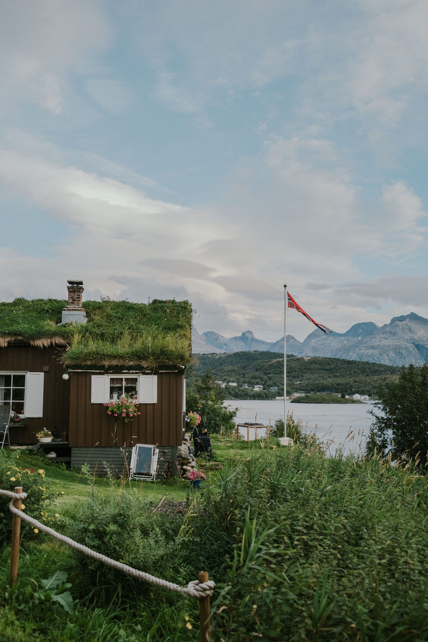Brown wooden cabin with traditional grass roof and flag pole against mountainous Norwegian backdrop.