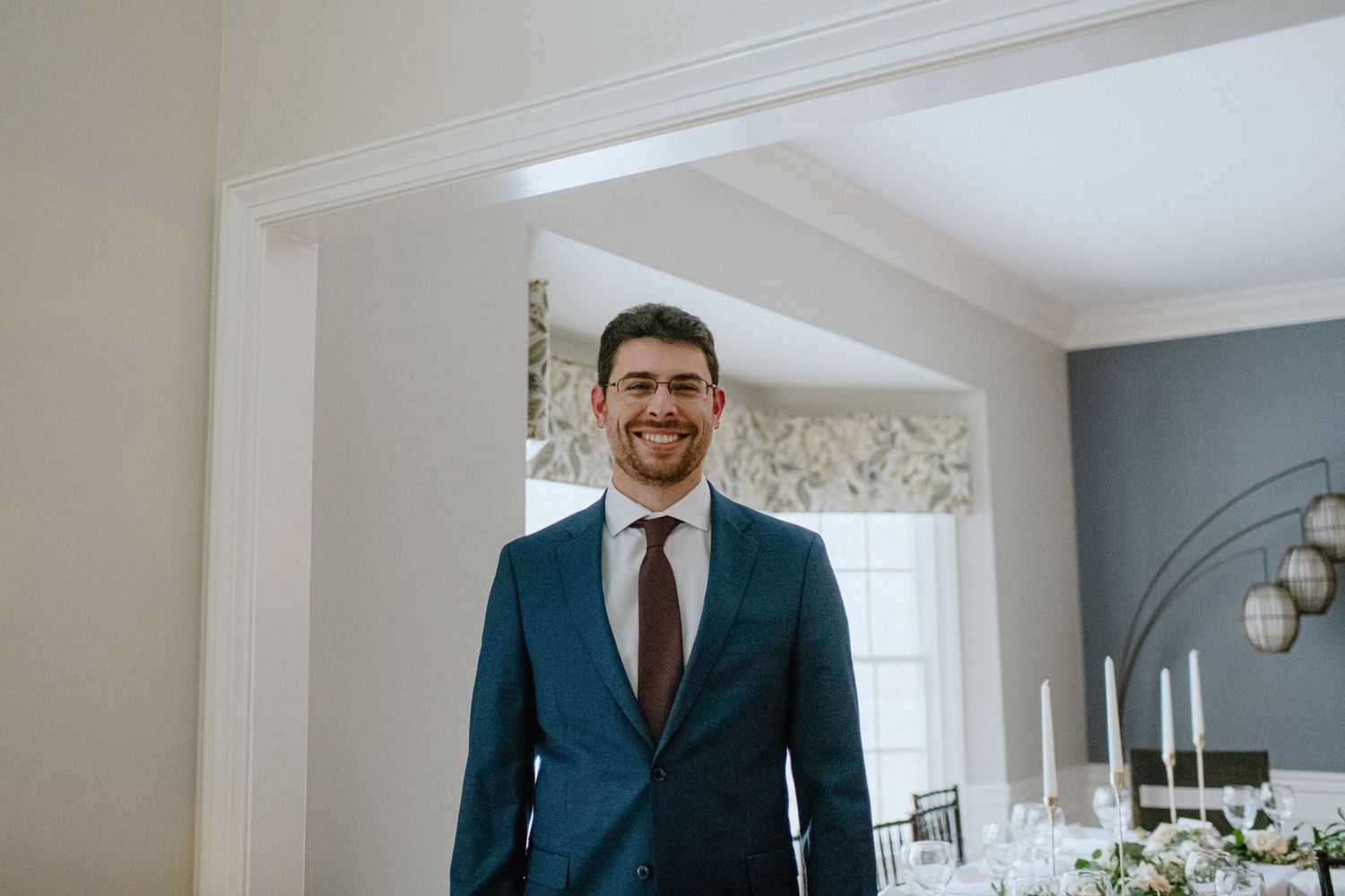 A groom in a dark teal suit and brown tie smiles warmly in an elegant indoor setting on his wedding day.