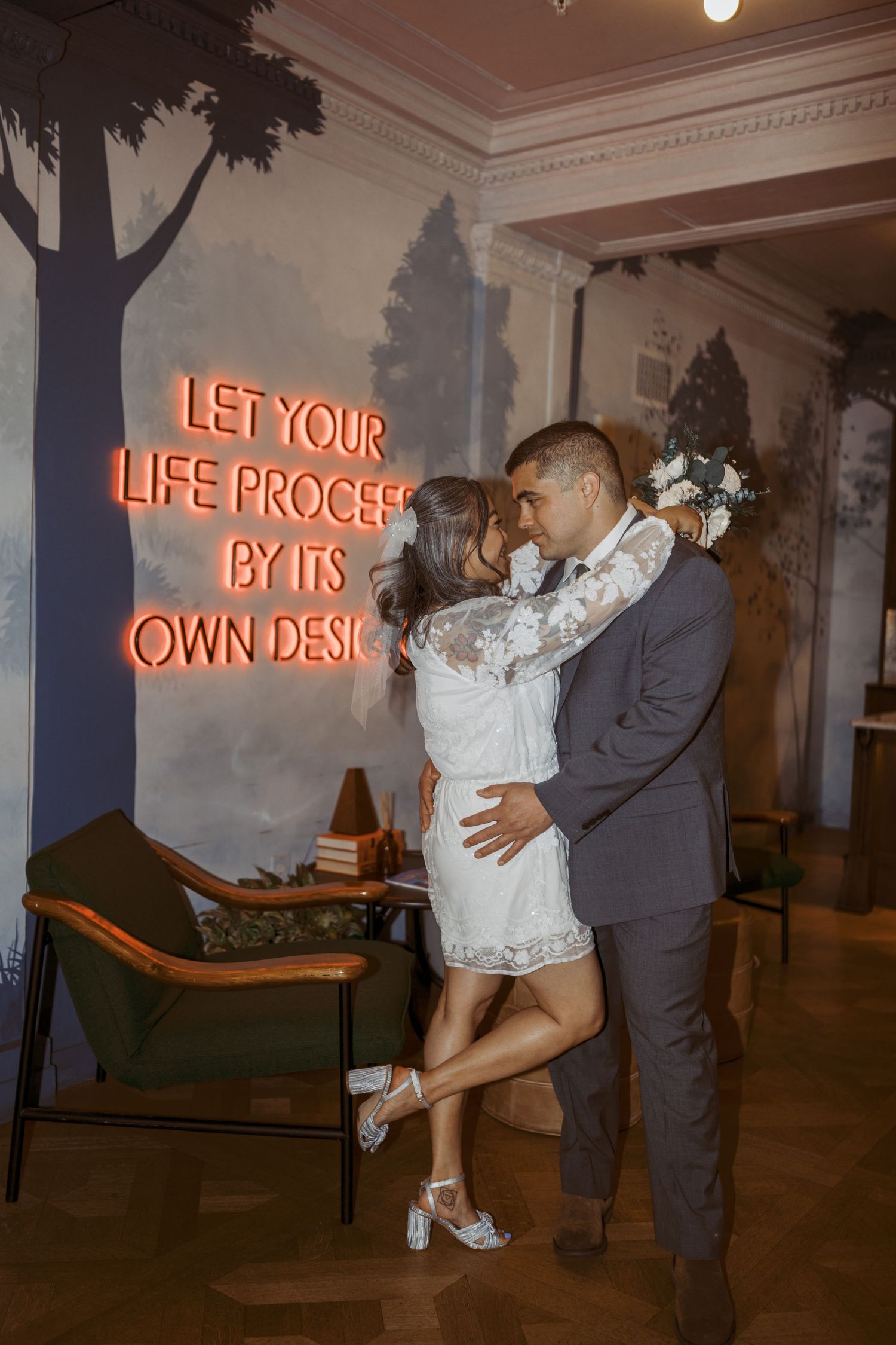 A couple embraces near a neon sign reading 'Let your life proceed by its own design'.