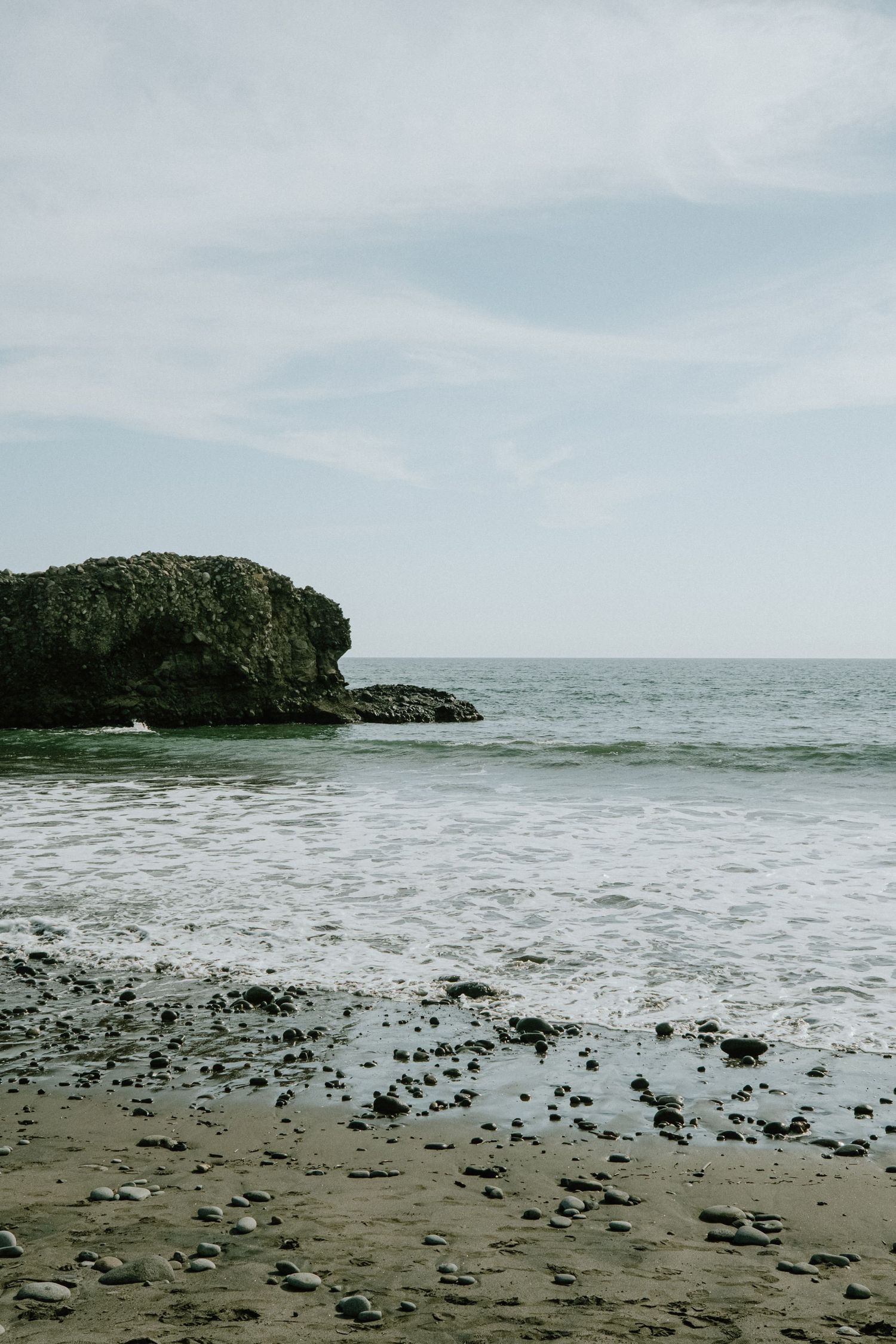 Rocky coastline with waves crashing against a large rock formation on a cloudy day.