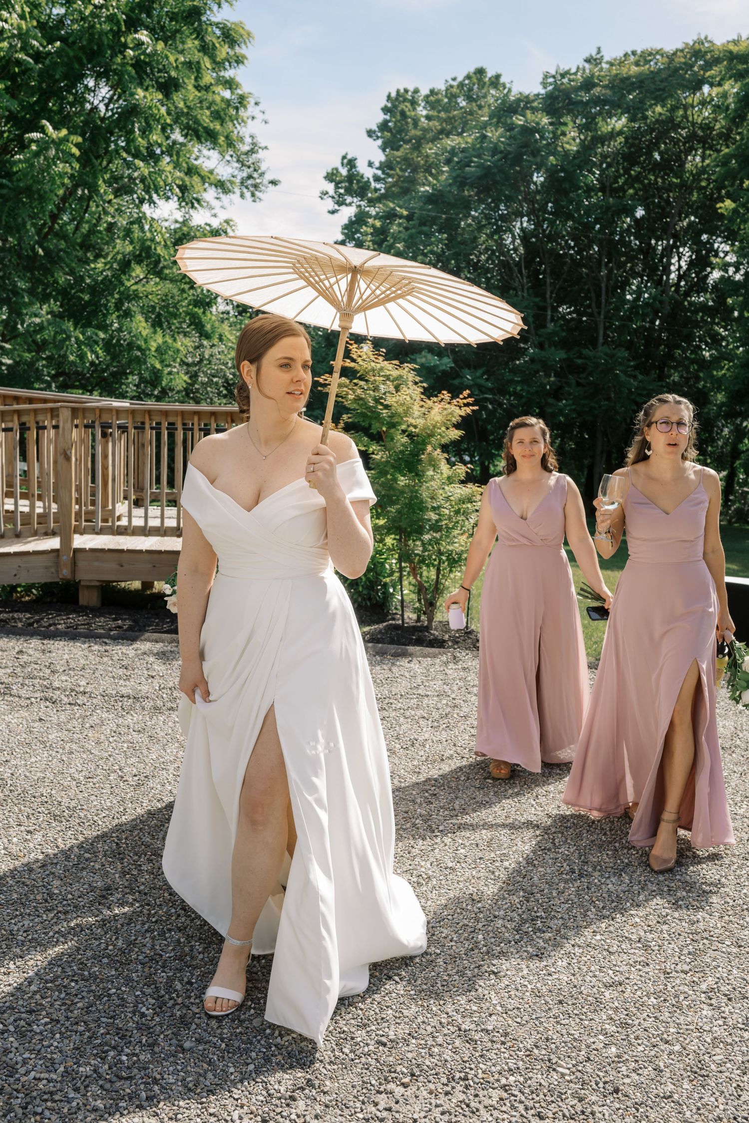 Bride in white gown with slit holding parasol walks with bridesmaids in pink dresses on gravel path.