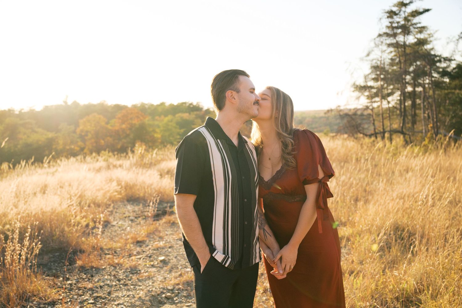 A couple shares a romantic moment in a sunlit golden field during sunset with evergreen trees in the background.