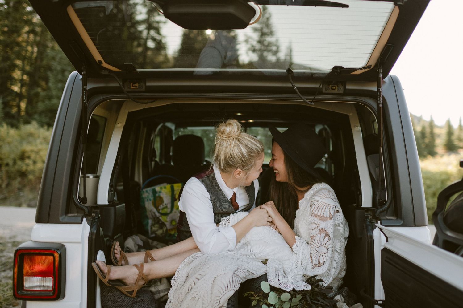 A couple sits together in the back of a Jeep with the trunk open in a forest setting.