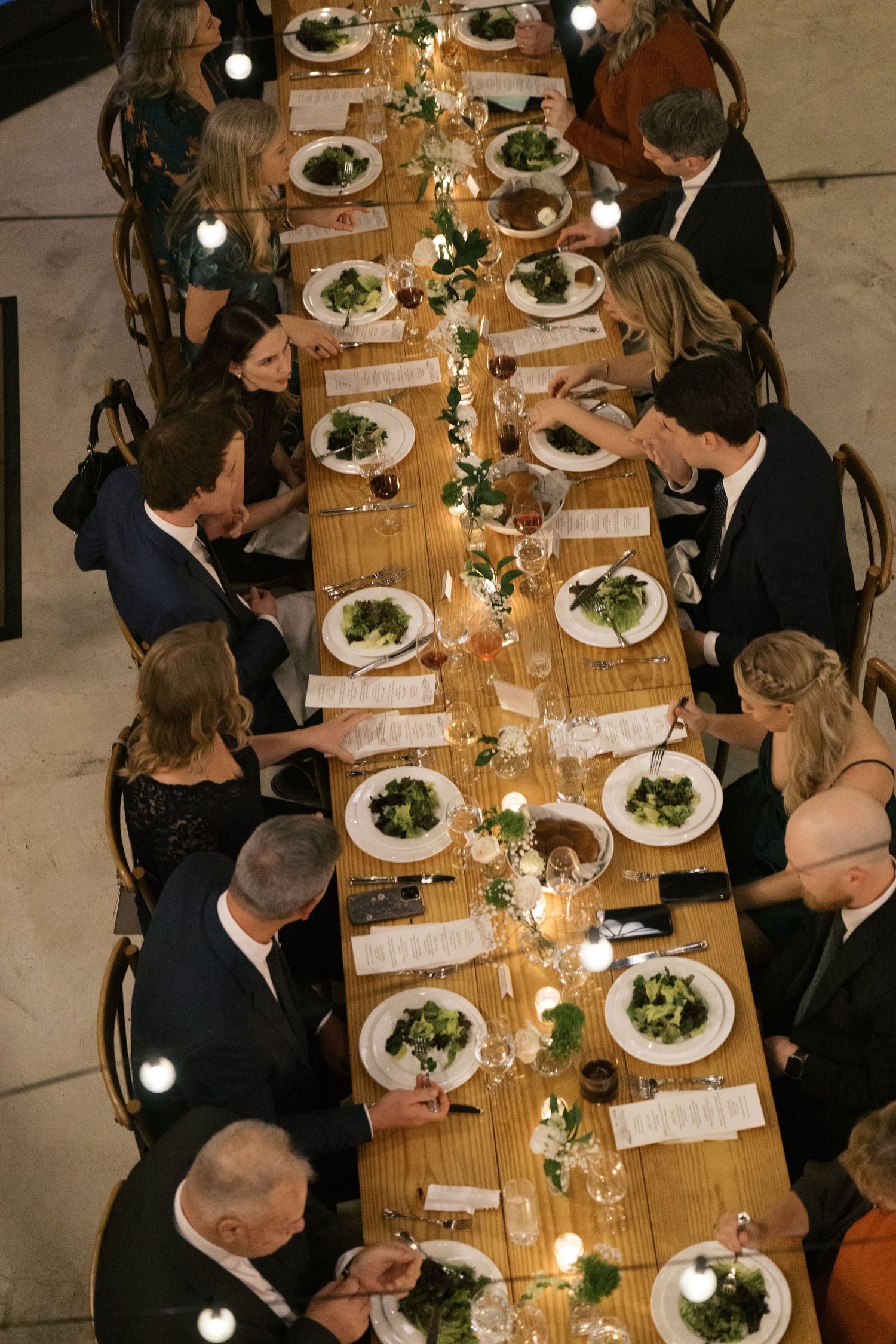 Overhead view of elegant wedding reception dinner with long wooden table, white plates and floral centerpiece decorations.