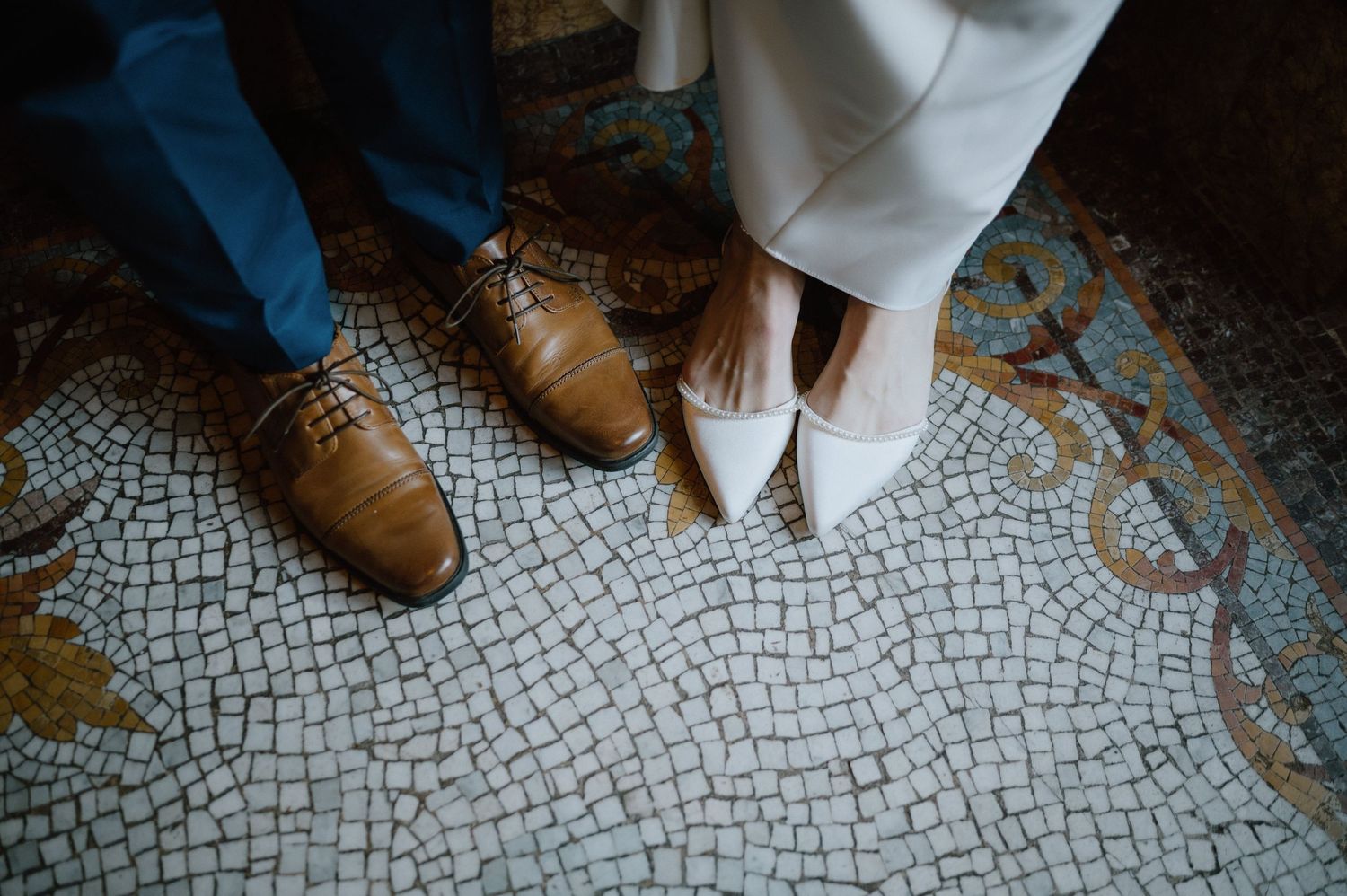 Close-up of contrasting footwear - brown dress shoes and white heels on mosaic tile floor.