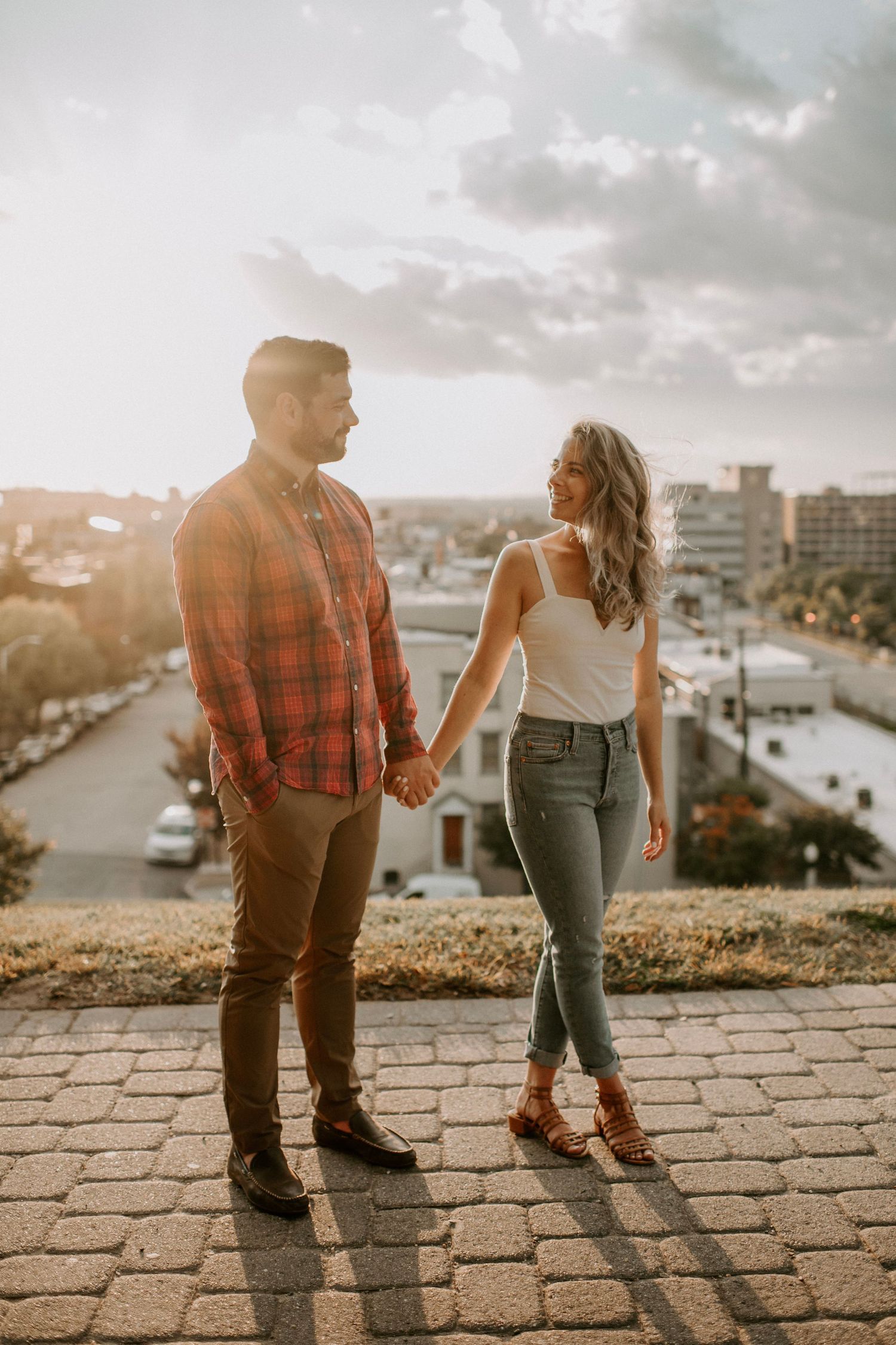Couple standing hand in hand on stone path overlooking urban landscape at sunset.