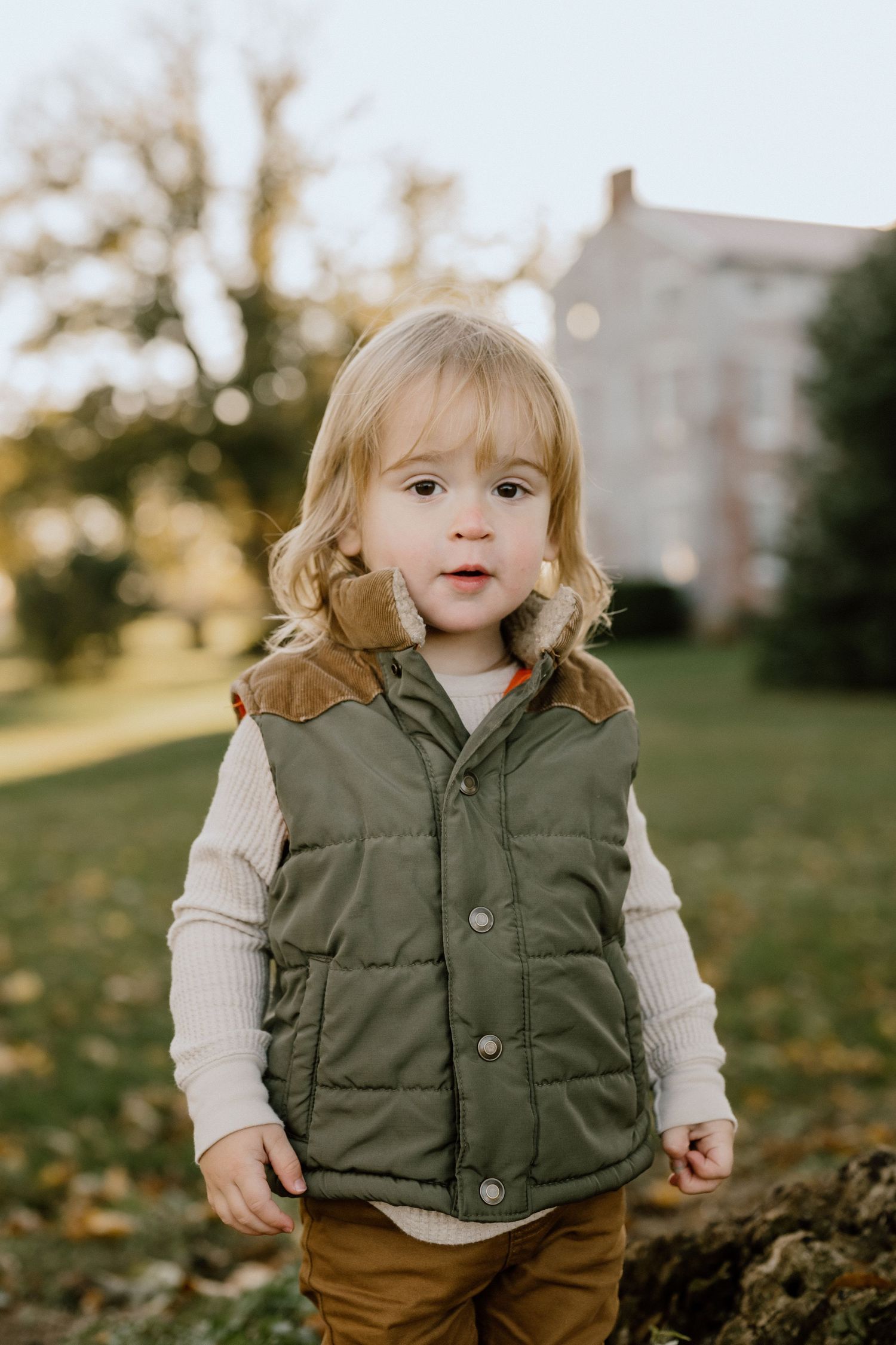 Child in olive green puffy vest and cream sweater stands in autumn yard.