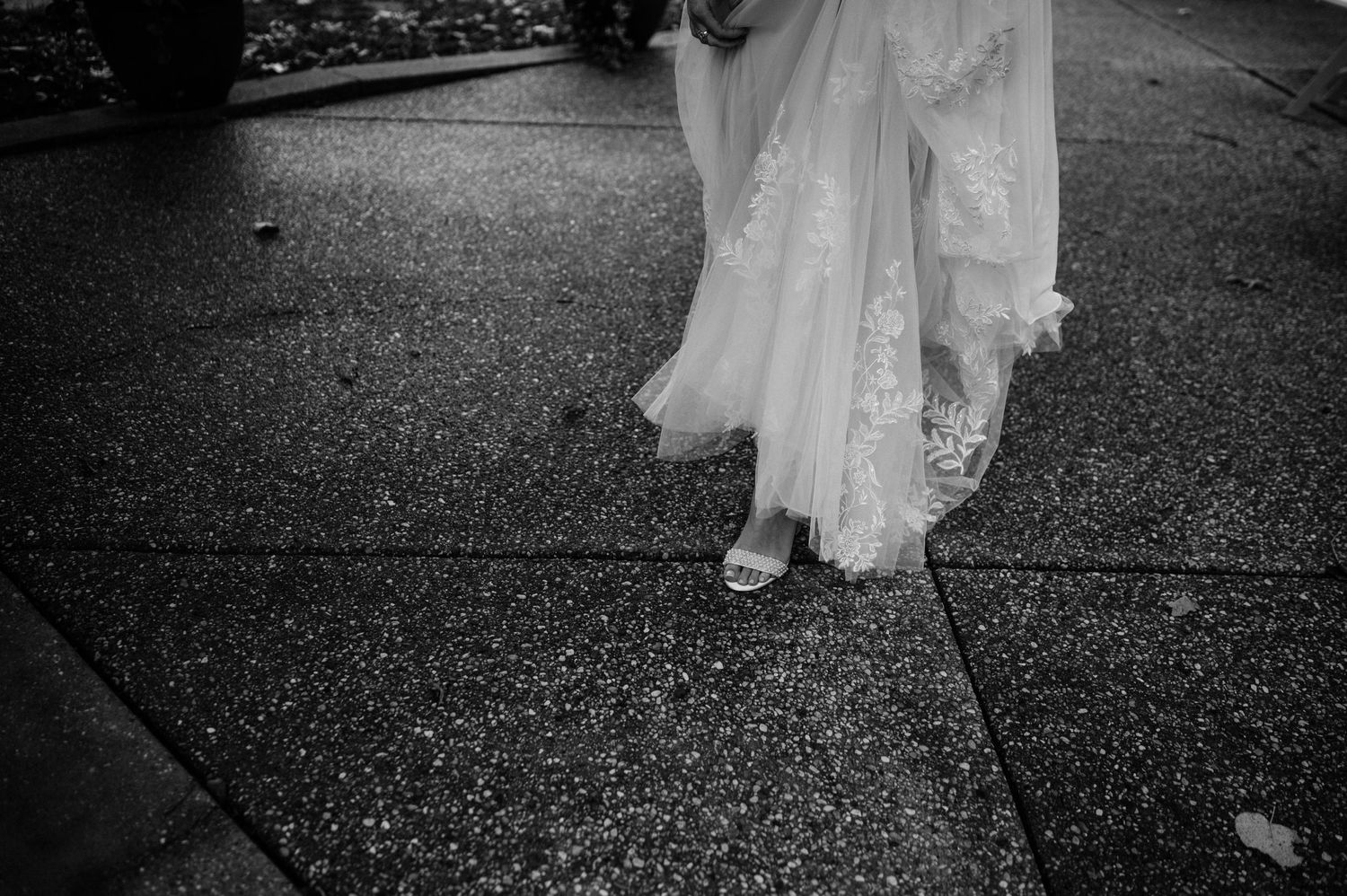 Black and white detail shot of bride's dress hem and shoes on textured ground.