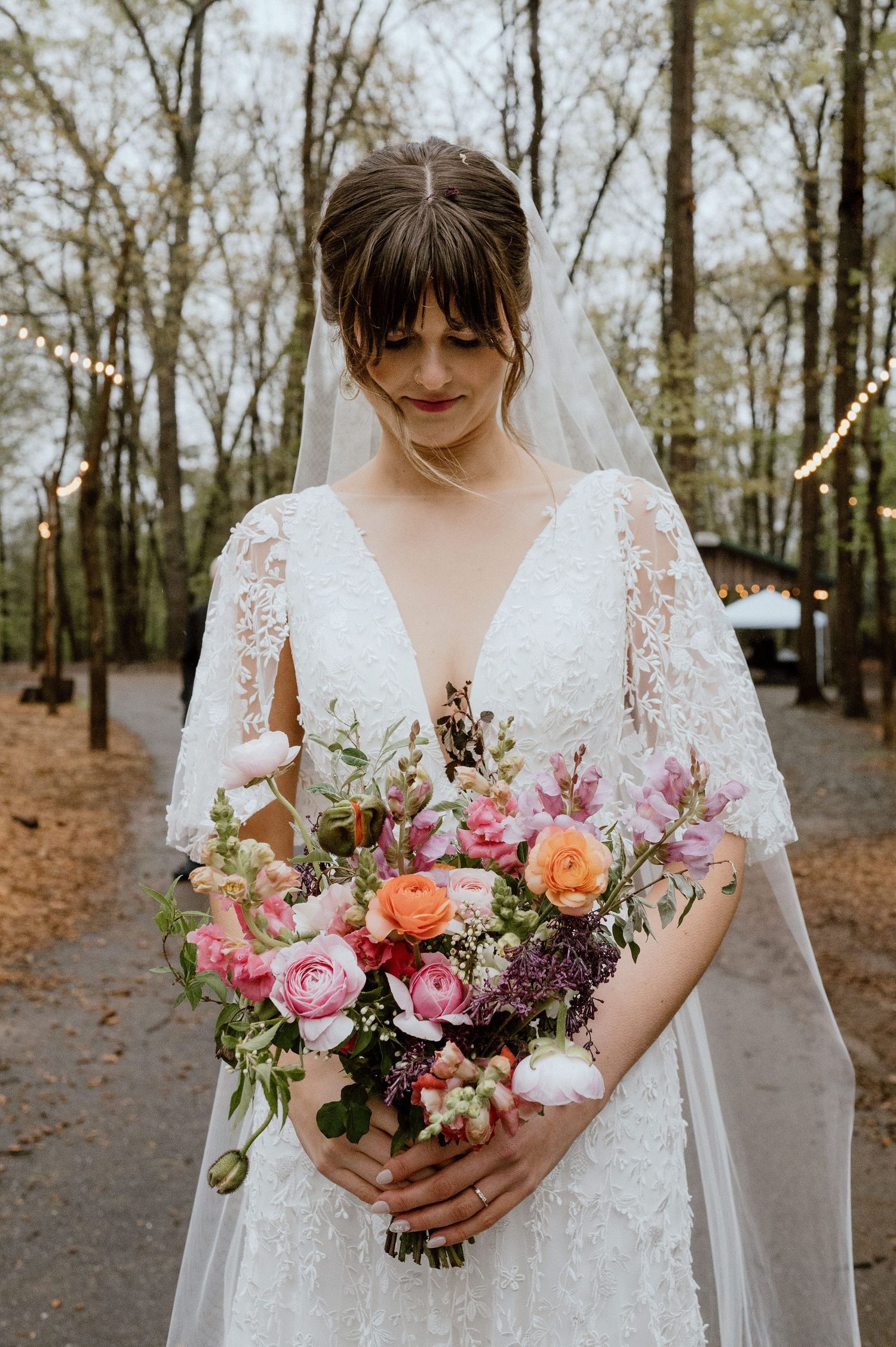 A bride holds a stunning bouquet of mixed flowers in pink, coral and purple against her lace wedding dress.