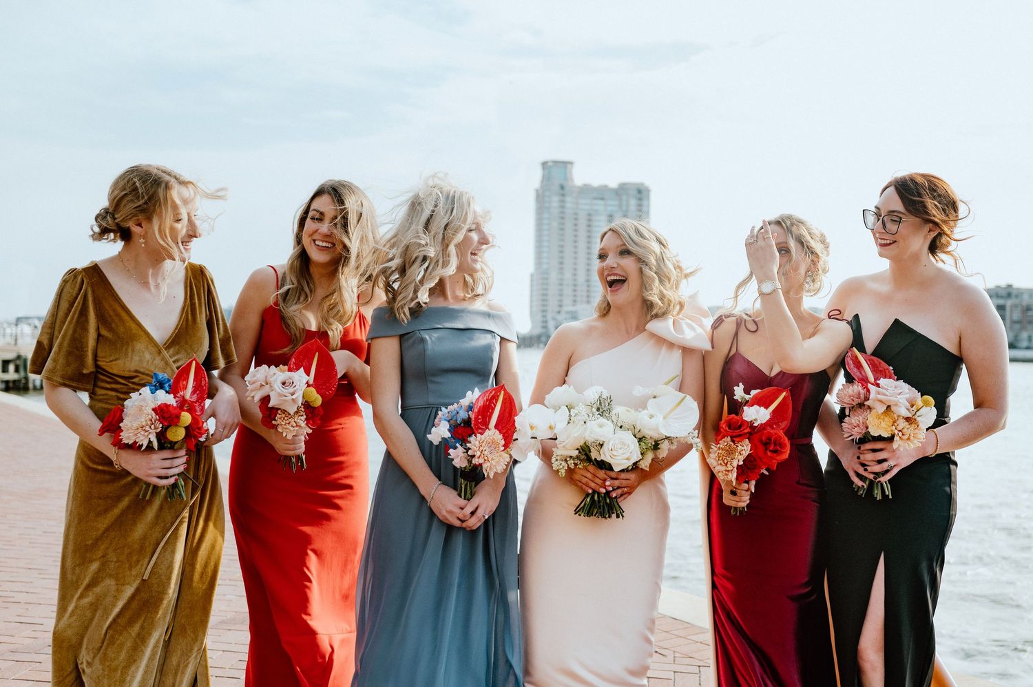 Bridesmaids in mix-matched formal dresses holding bouquets pose together on waterfront.