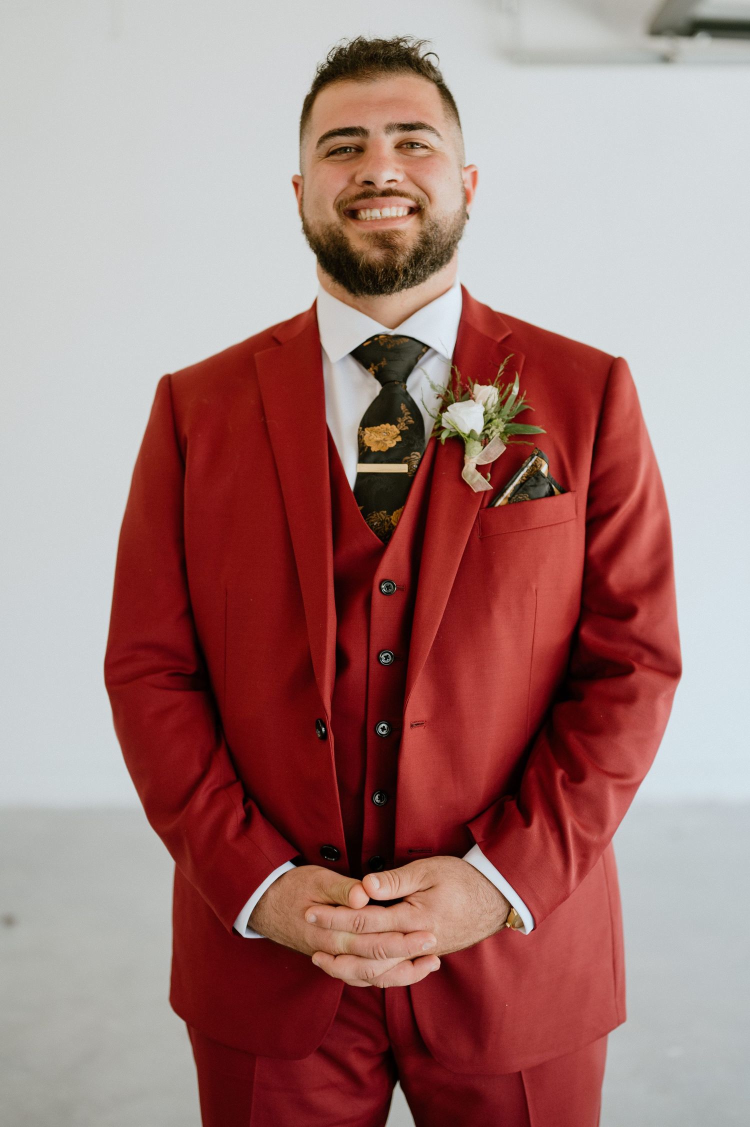 A groom in a crimson suit with boutonniere smiles warmly against a white backdrop.