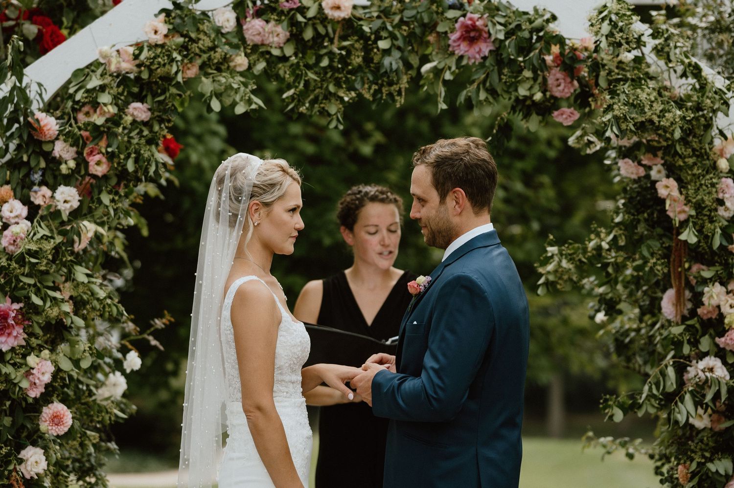 Wedding ceremony under a floral arch with pink roses and white lattice in a garden setting.