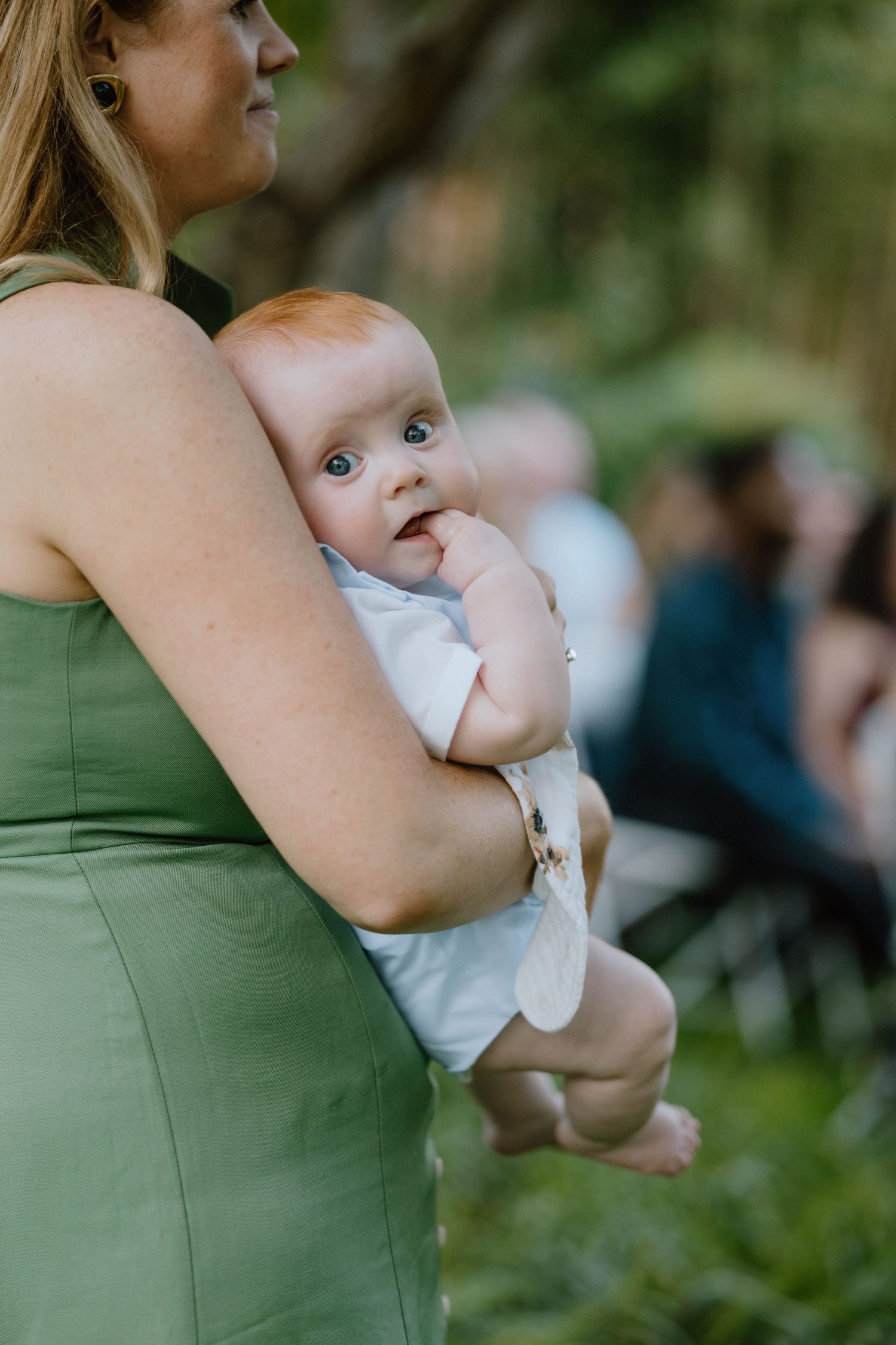 Baby in white outfit being held at an outdoor gathering.