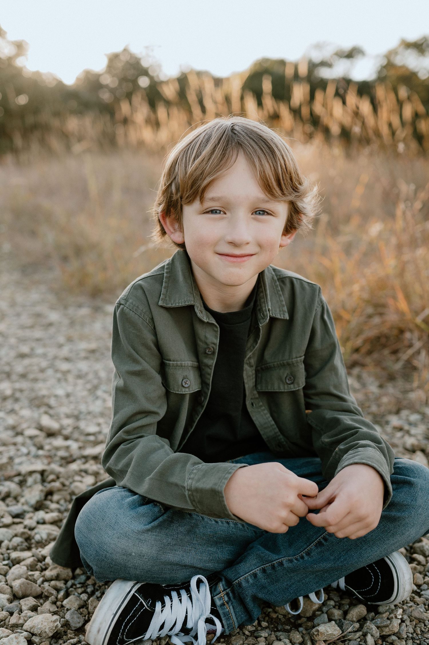 A child sits cross-legged on rocky ground wearing sneakers and a green jacket at sunset.