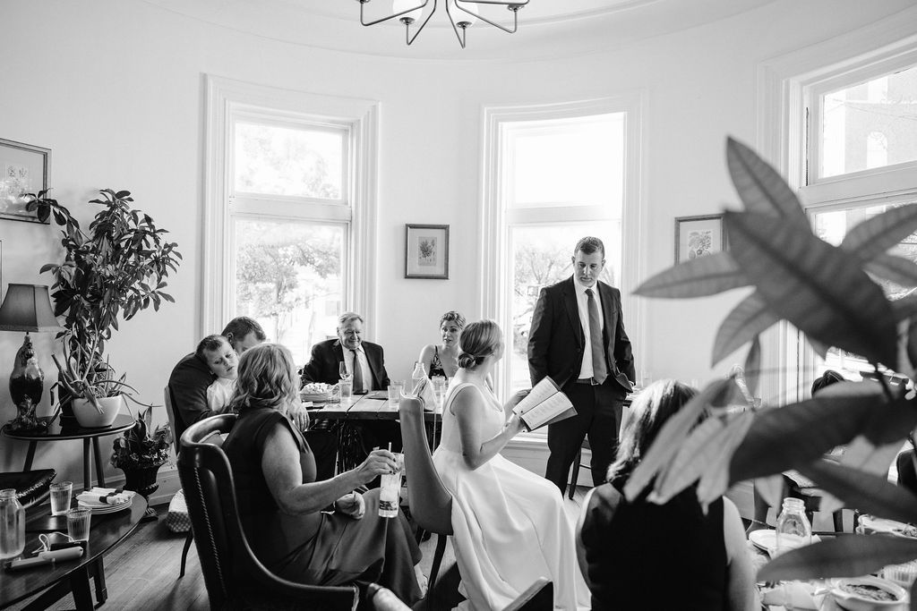A black and white photo of a wedding celebration in a bright room with large windows and indoor plants.