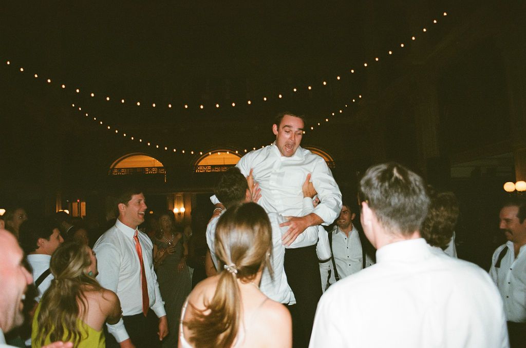 String lights illuminate a dark reception venue as guests dance below.
