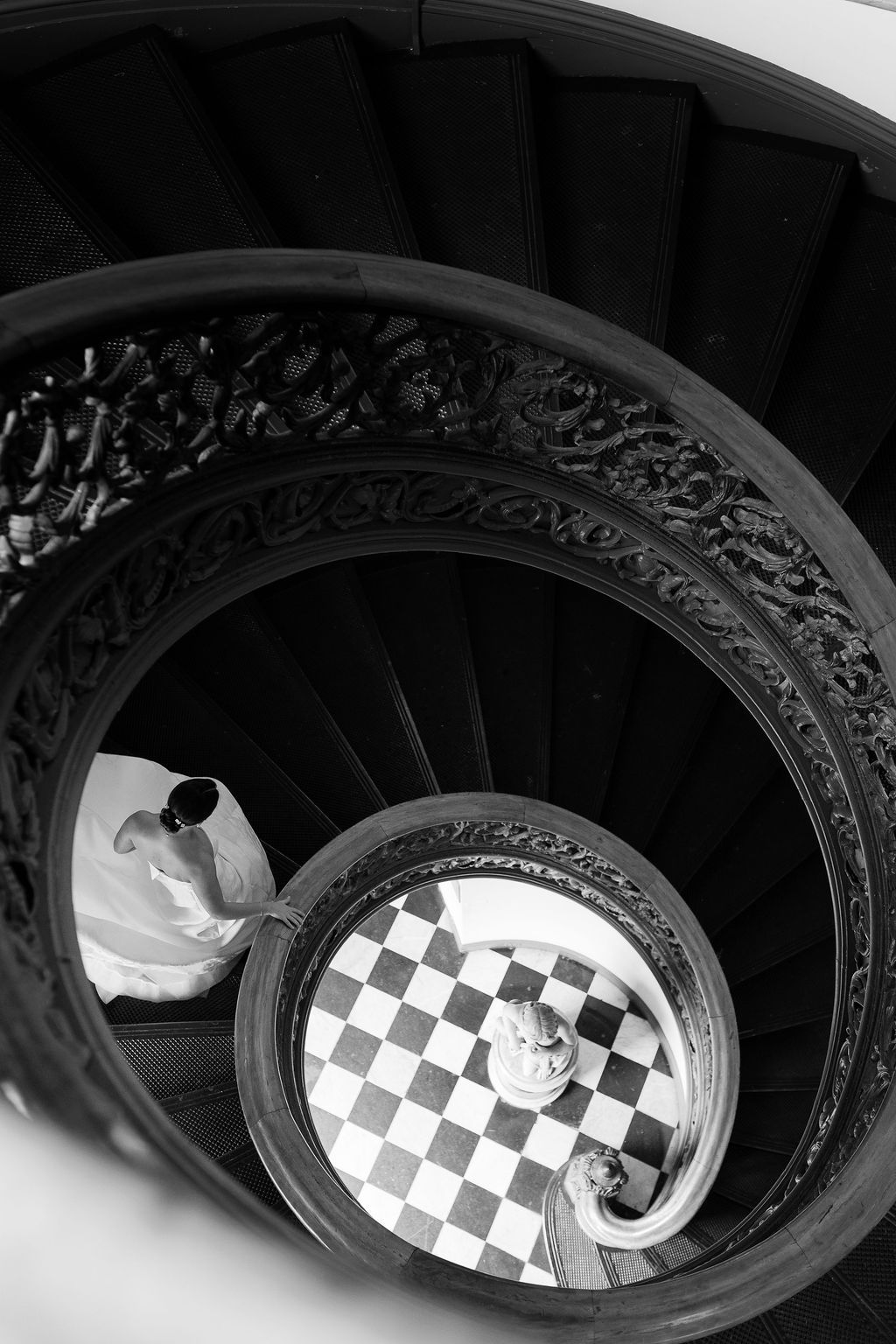 Spiral staircase with ornate railing frames couple on black and white checkered floor below.