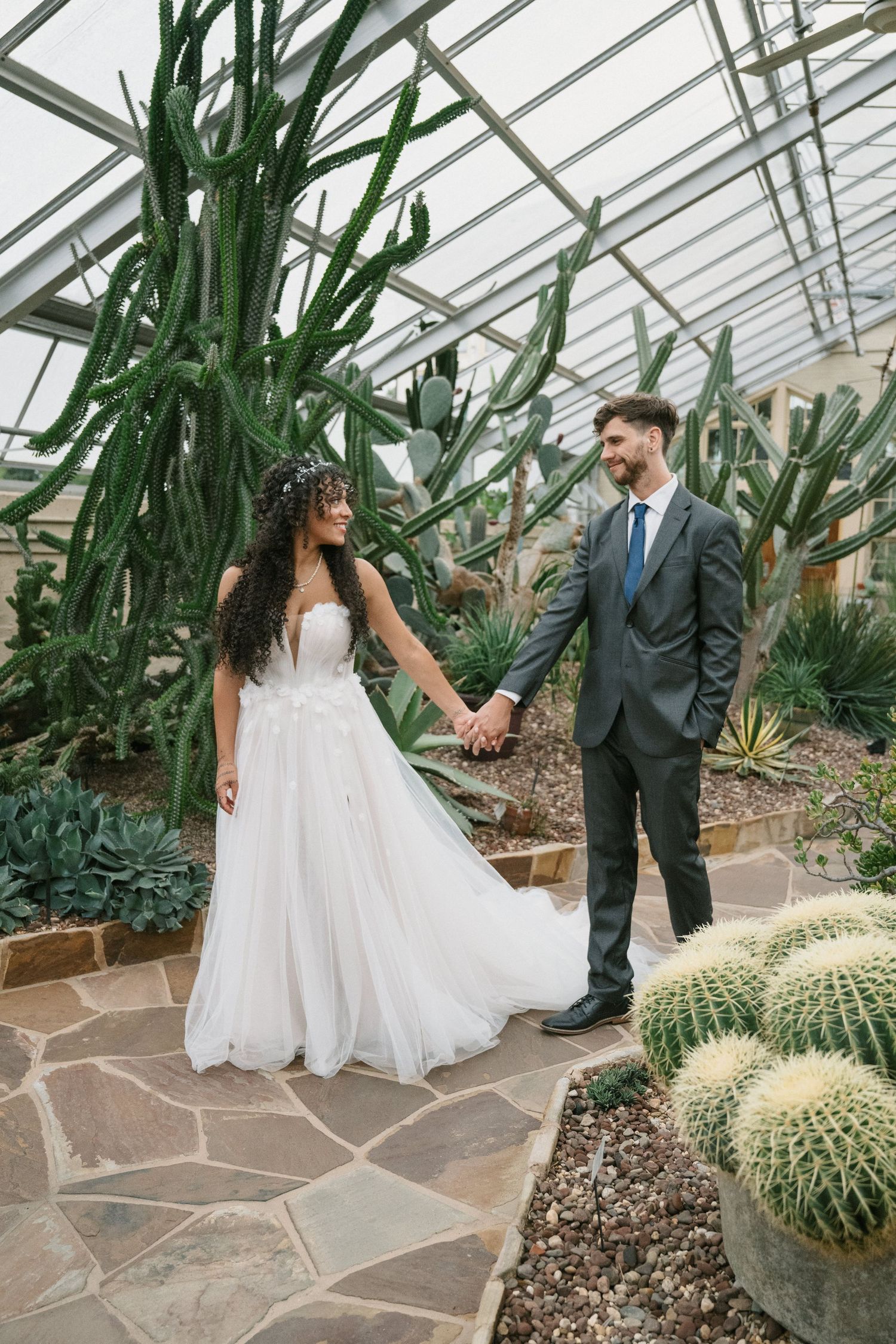 Bride and groom hold hands in a lush greenhouse wedding venue surrounded by towering cacti and succulents.