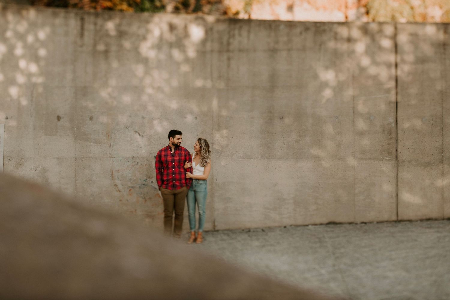 Couple standing together against a concrete wall outdoors, sharing a romantic moment in warm autumn light.