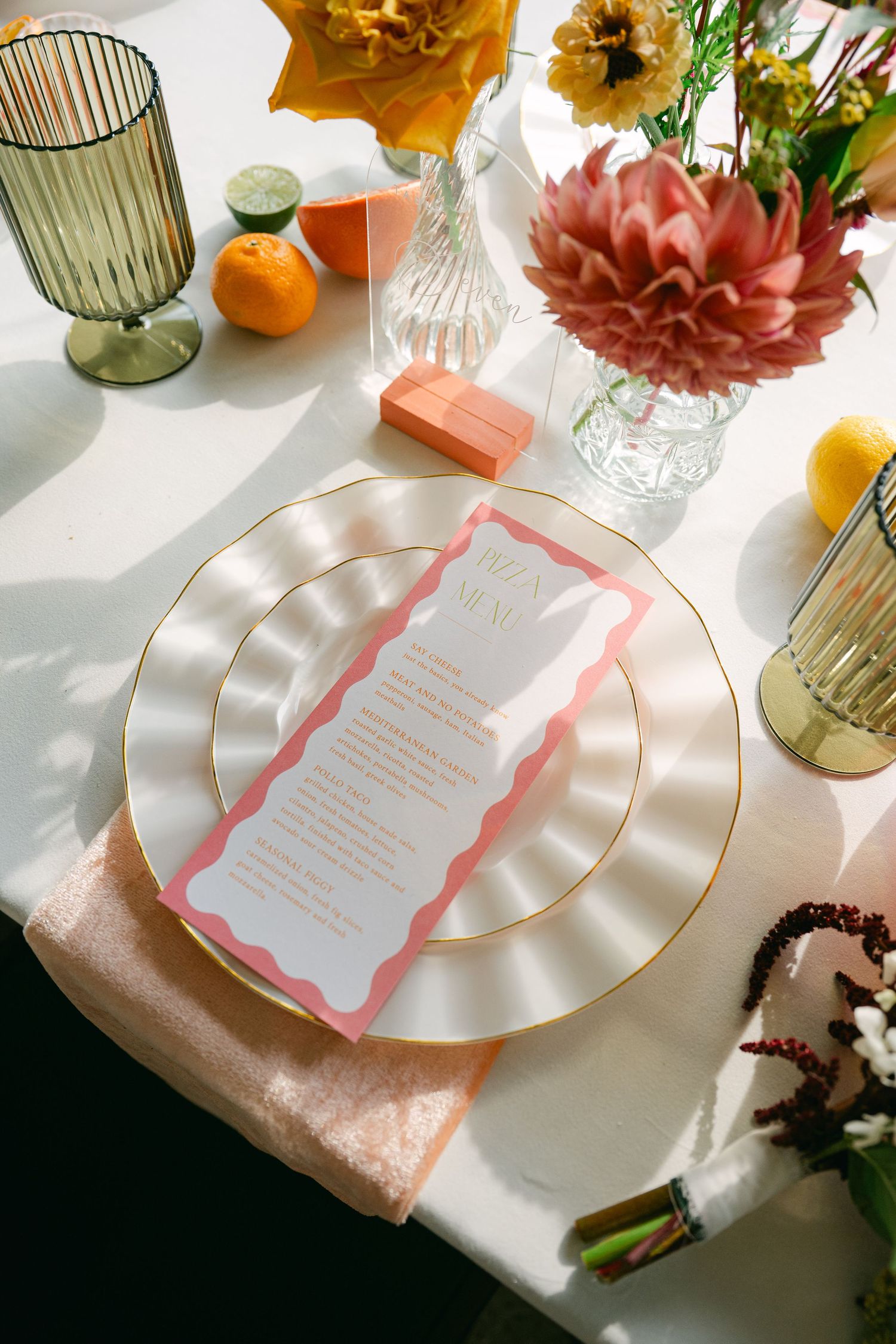 Elegant wedding table setting with pink menu card, gold-rimmed plate, colorful florals, citrus fruits, and sage green glassware.
