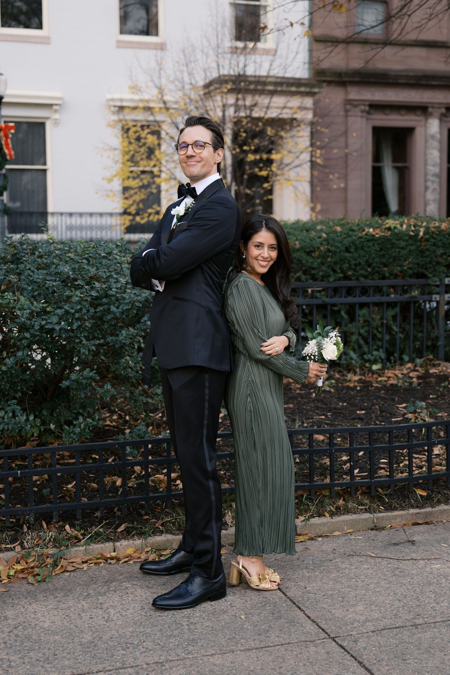 Autumn wedding portrait with couple posing back-to-back on sidewalk near brownstone buildings.