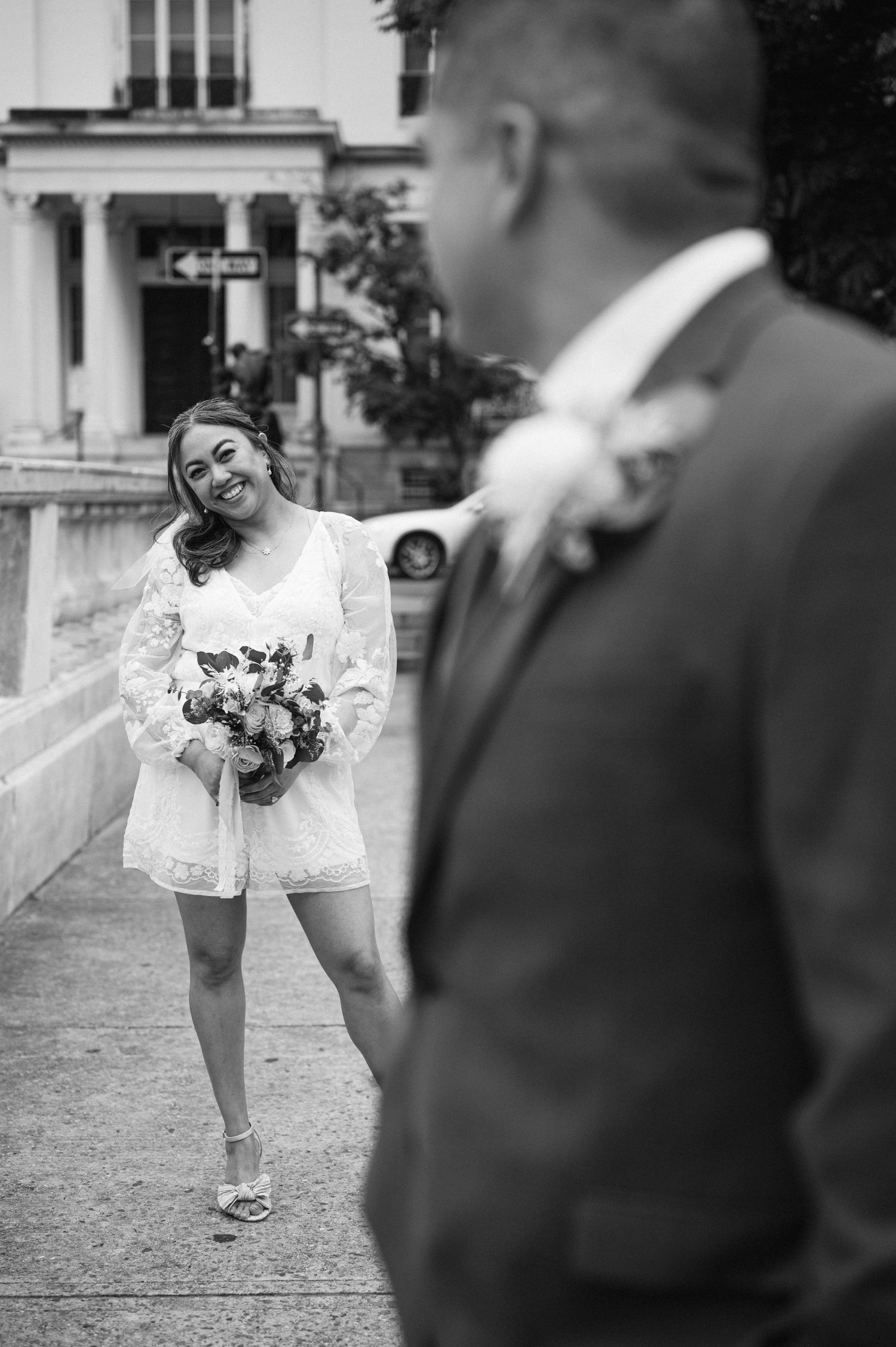 A black and white wedding photo shows a bride in a short white dress holding a bouquet as she smiles at her groom outdoors.