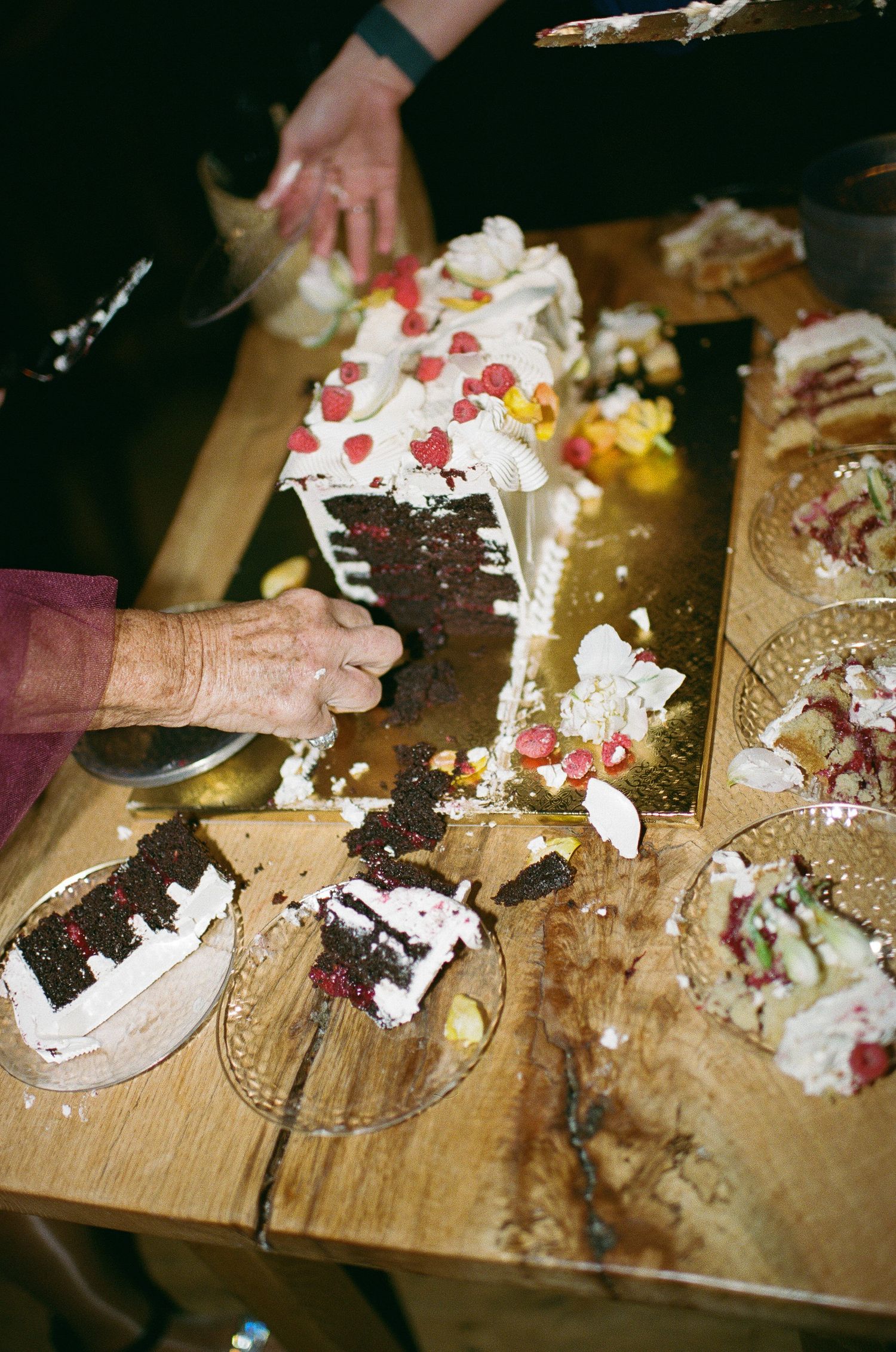 Hands cutting and serving slices of decorated chocolate cake with whipped cream and berries on a wooden cutting board.