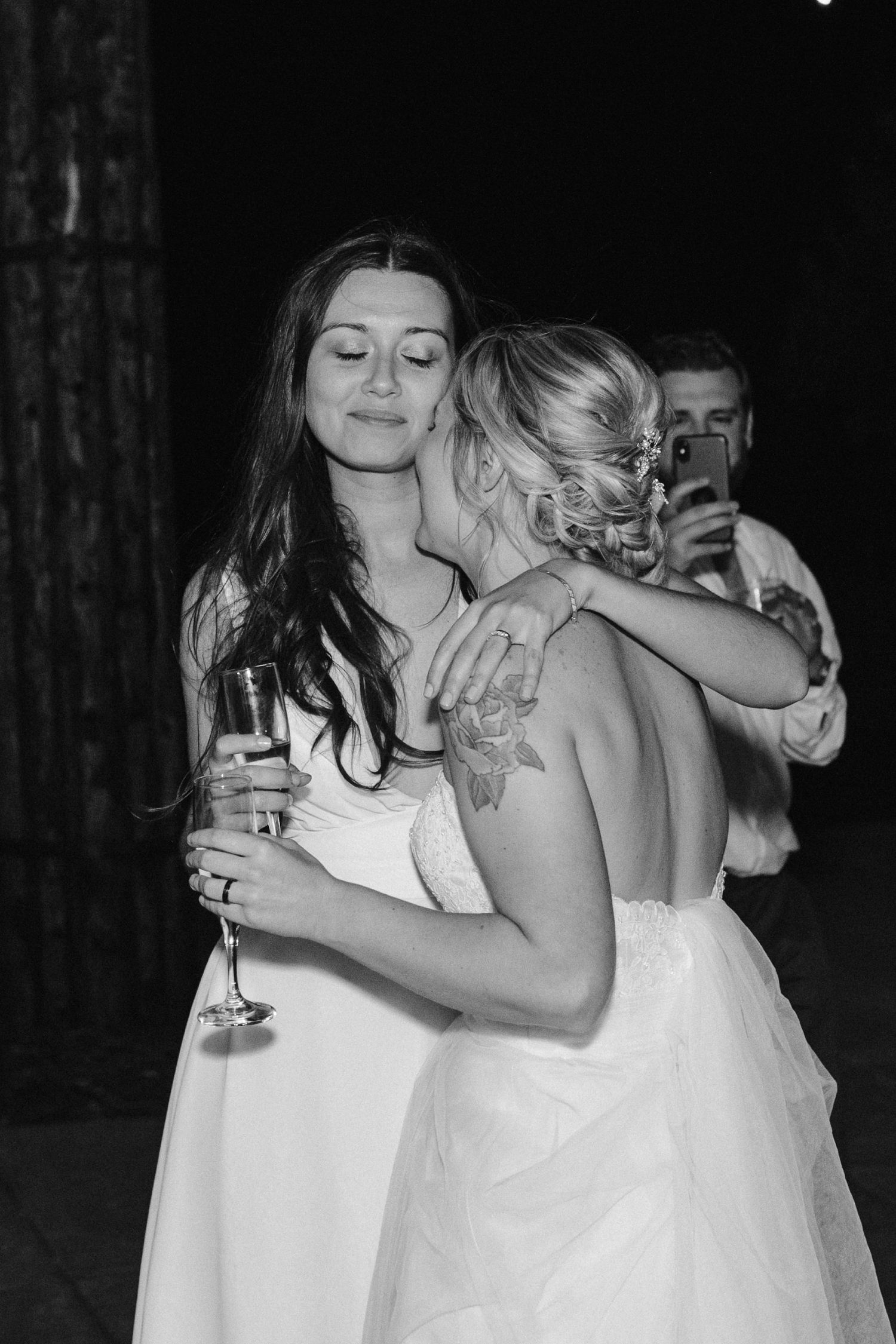 Black and white photo capturing an intimate embrace between two people in white dresses at a celebration.