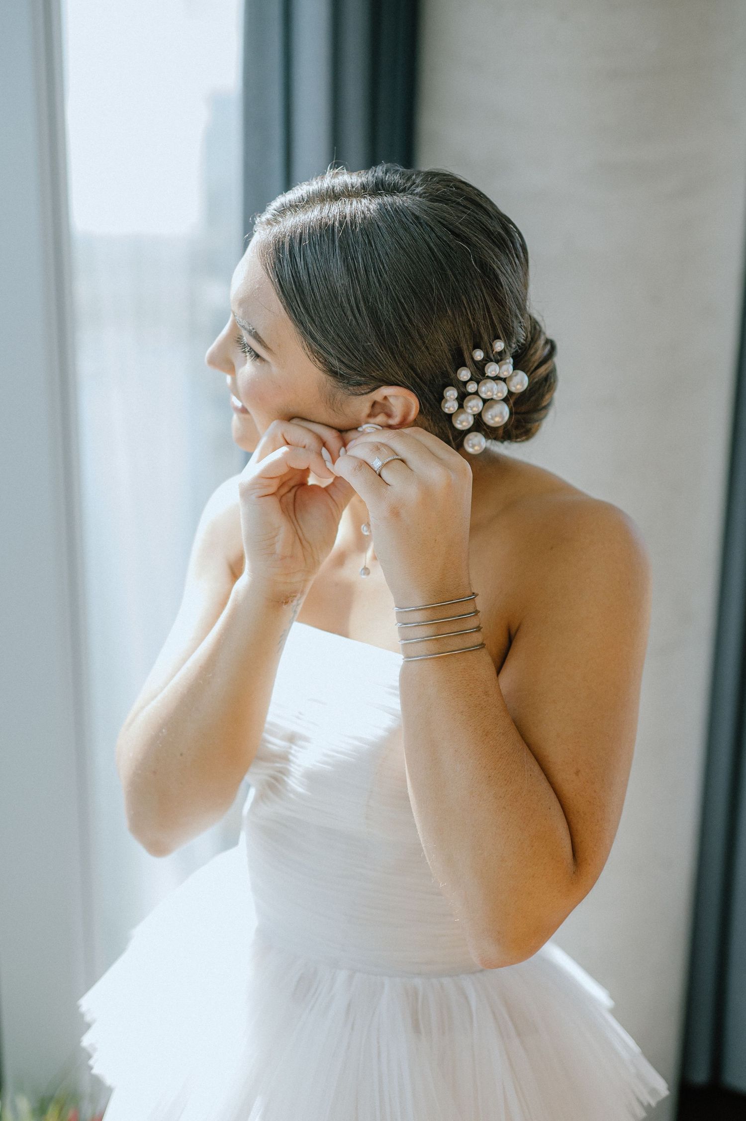 A bride puts on jewelry while getting ready, wearing an elegant white gown with delicate hair accessories.