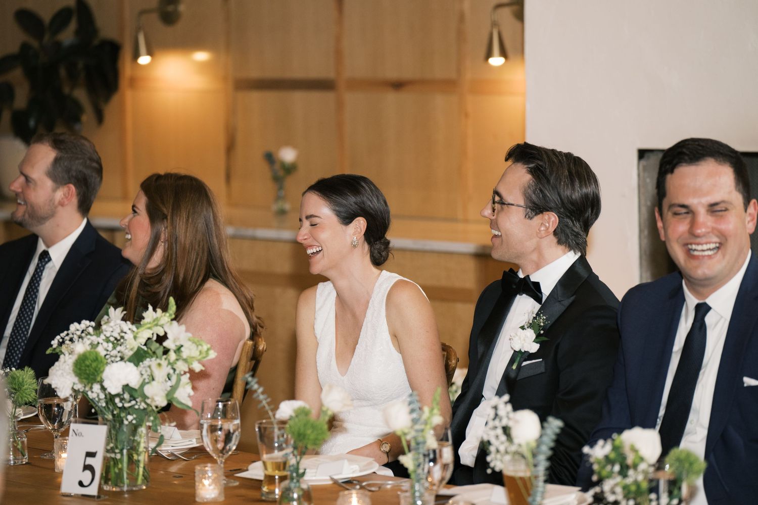 Wedding reception guests seated at a table with white floral centerpieces during dinner service at an indoor venue.