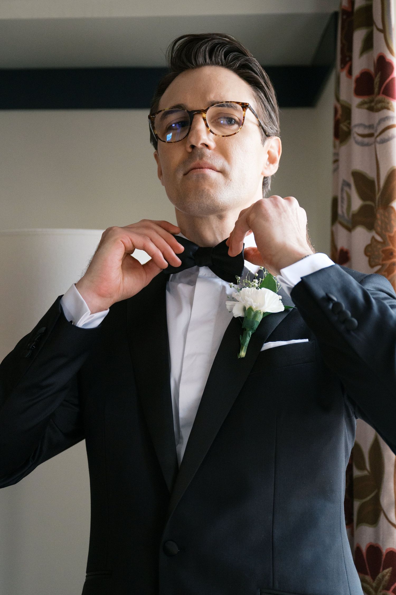 A groom in a black tuxedo and glasses adjusts his bow tie while wearing a white boutonniere.