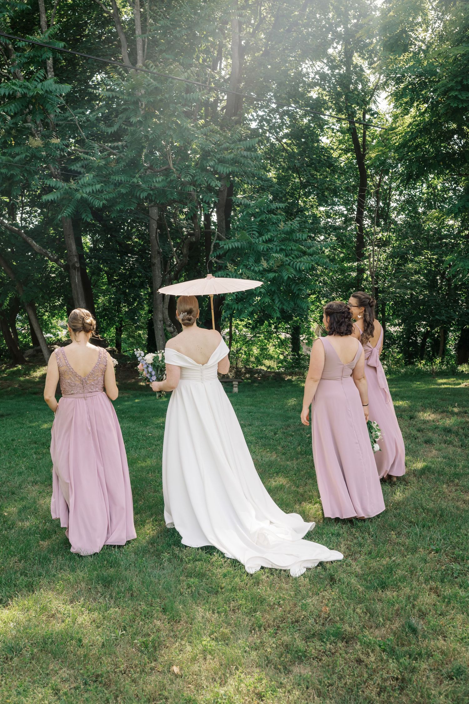 Wedding party walking on grass with bridesmaids in pink dresses and bride in white gown holding parasol.