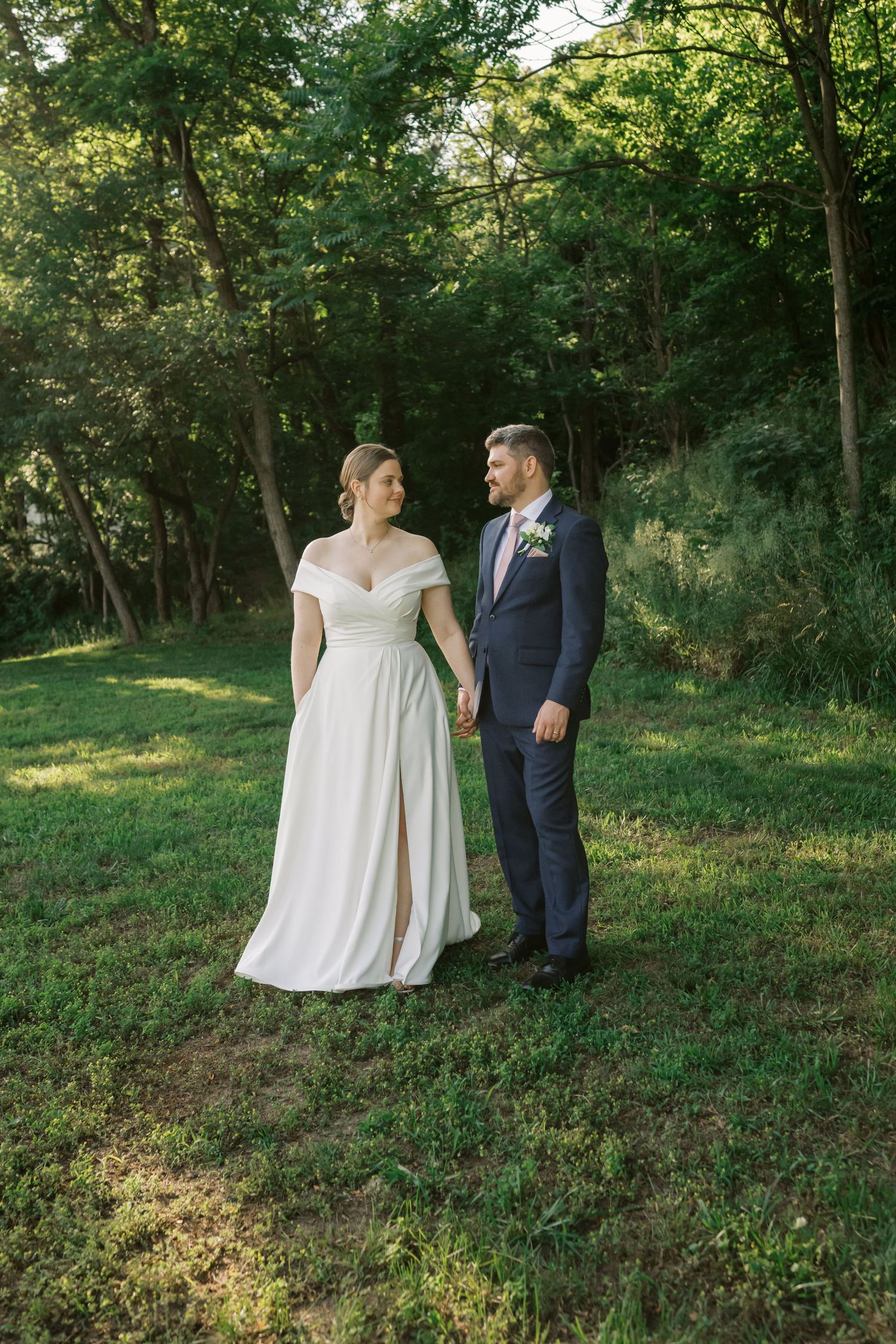 A couple in wedding attire poses together on a grassy lawn with trees in the background.
