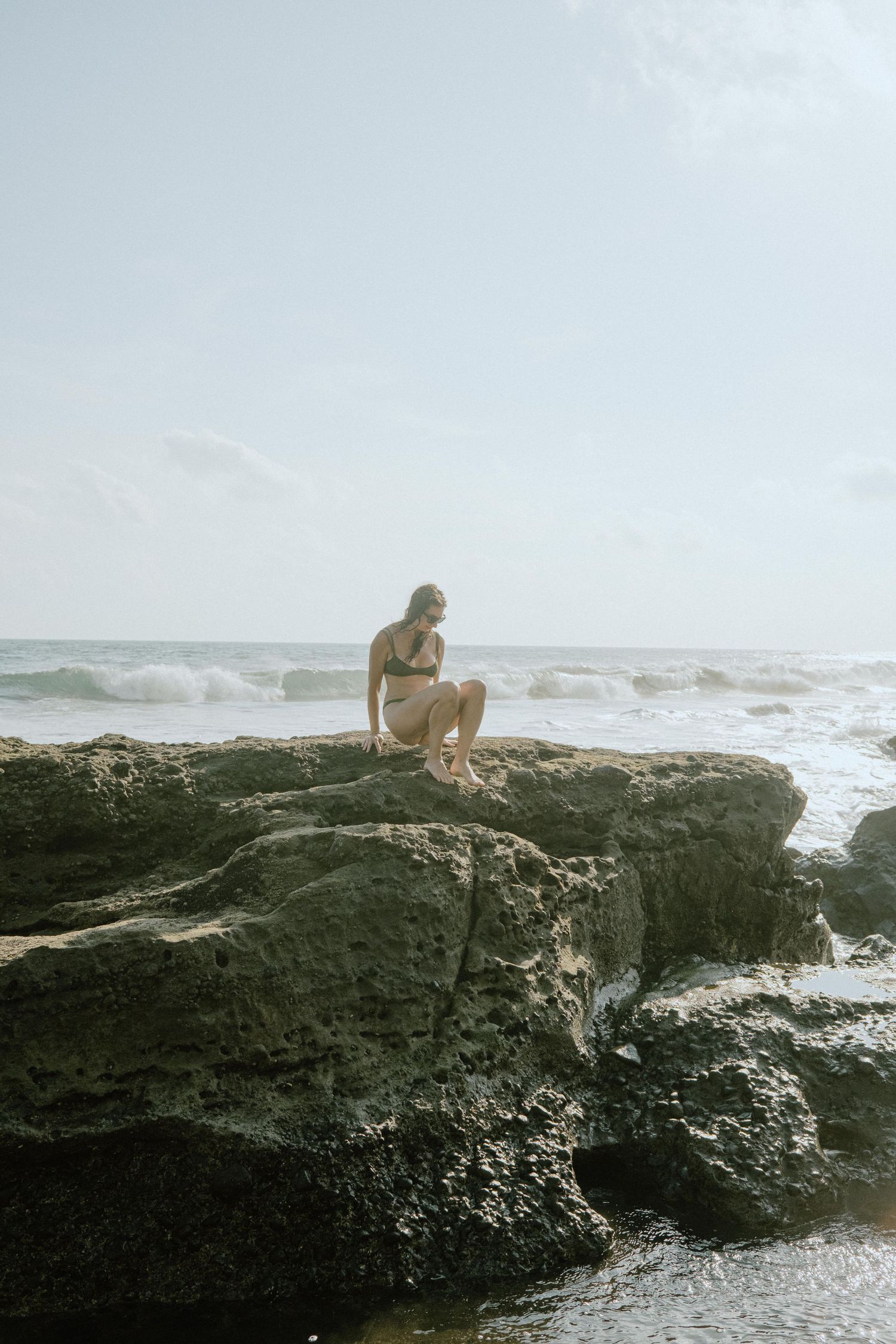 Person sitting on rocky coastal cliff overlooking ocean waves at sunset.