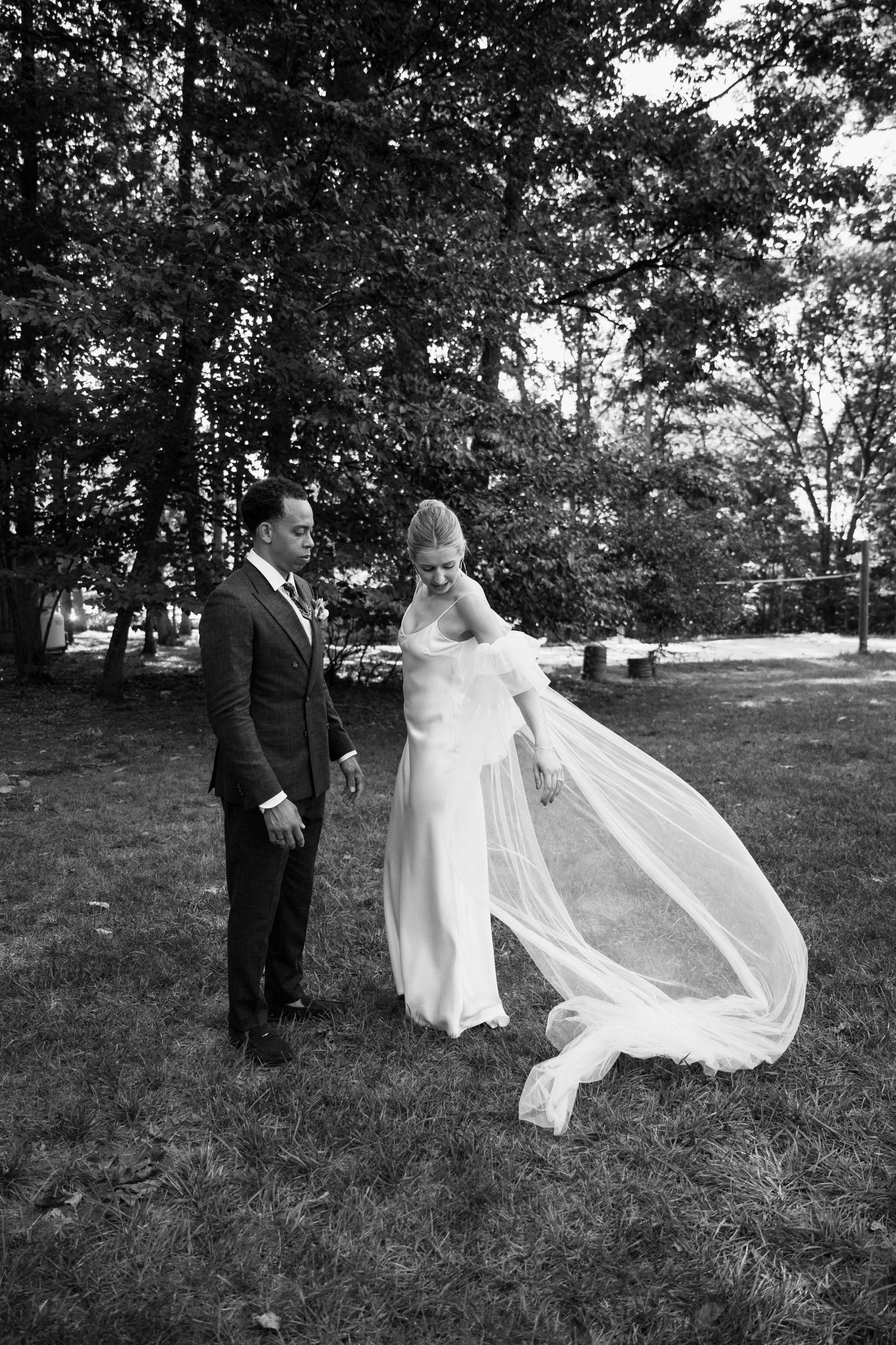 A black and white photo of newlyweds as the bride's veil catches the wind in a garden setting.