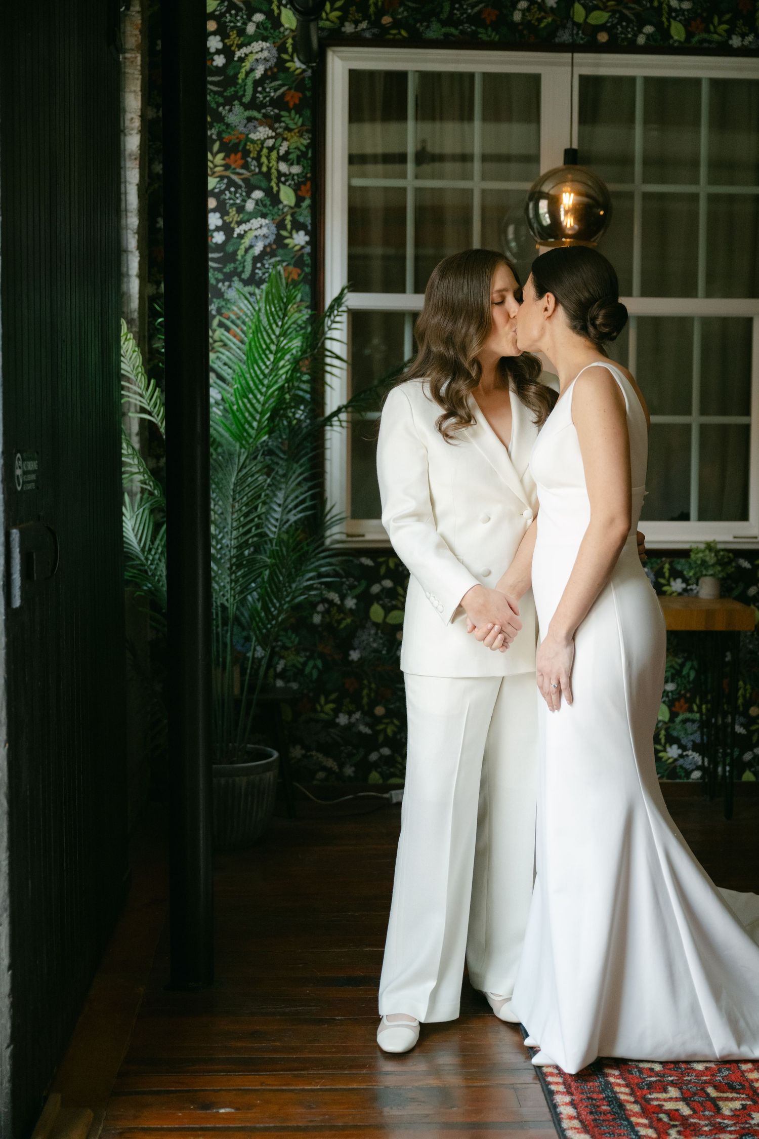Two people in white outfits share an intimate moment in a room with greenery and windows.