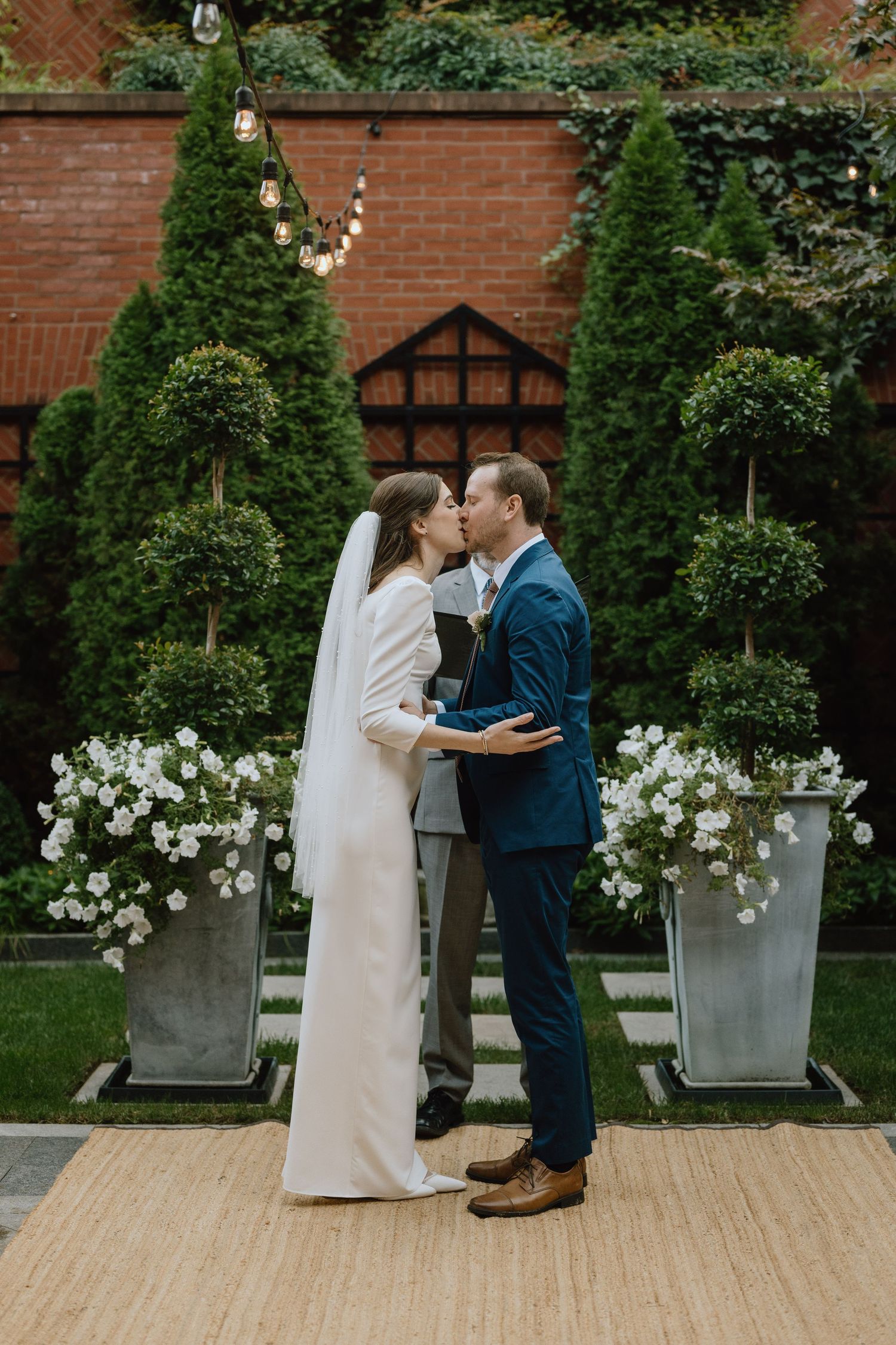 Elegant wedding ceremony scene with evergreen trees and white floral arrangements against a brick wall.