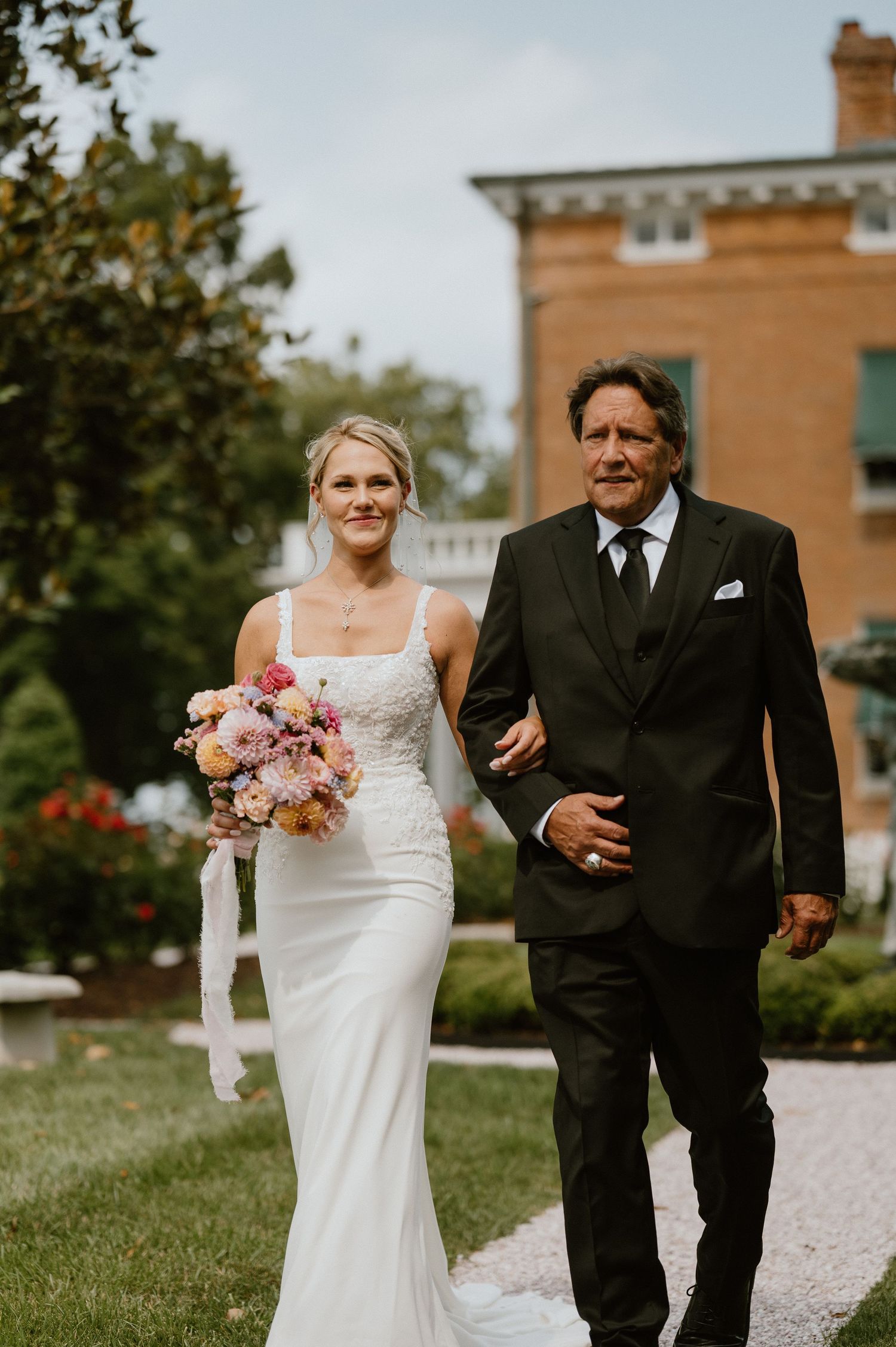 A bride in white gown carrying pink bouquet walks arm-in-arm with escort down garden path.