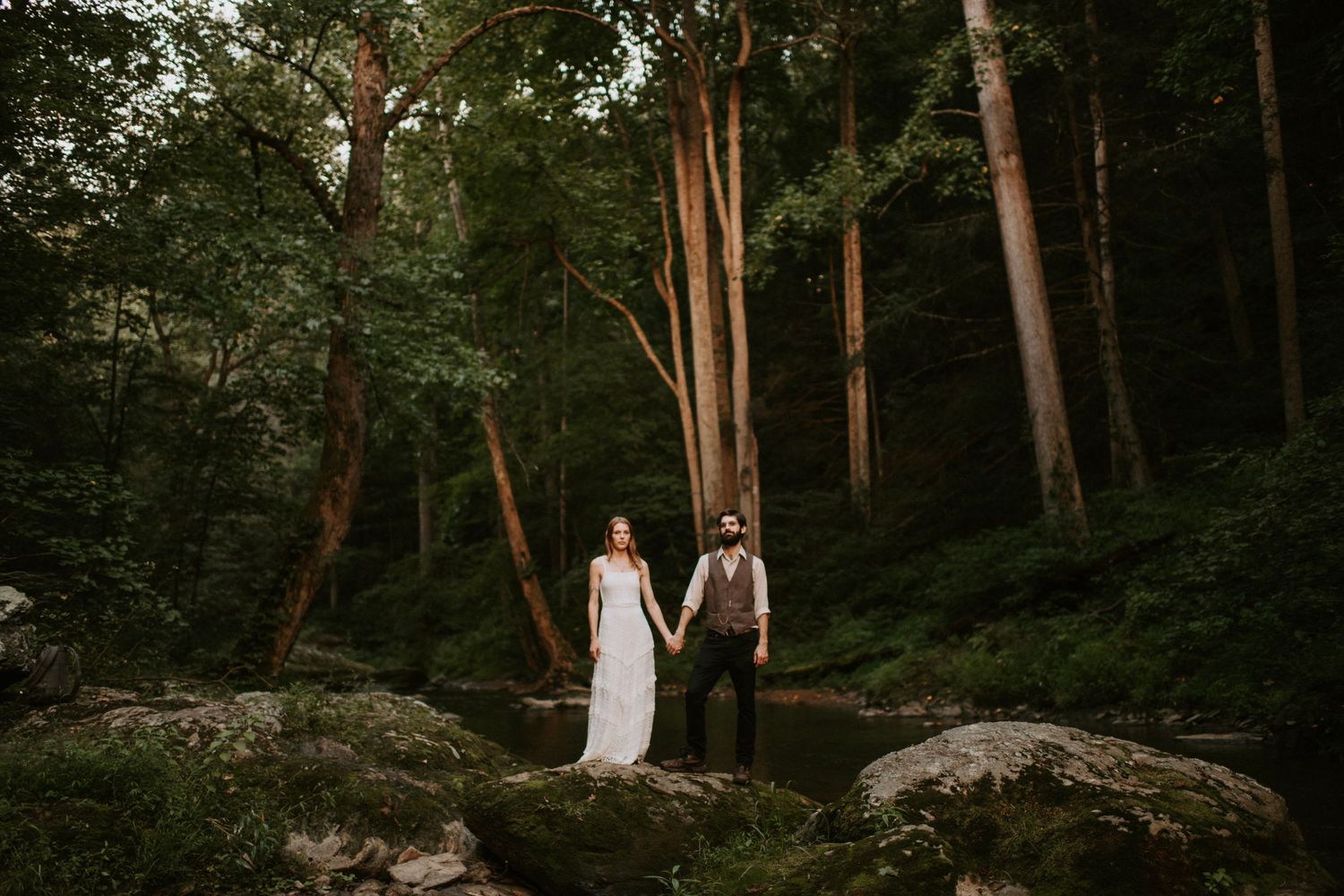 A couple stands on a large rock surrounded by tall sunlit trees in a forest.