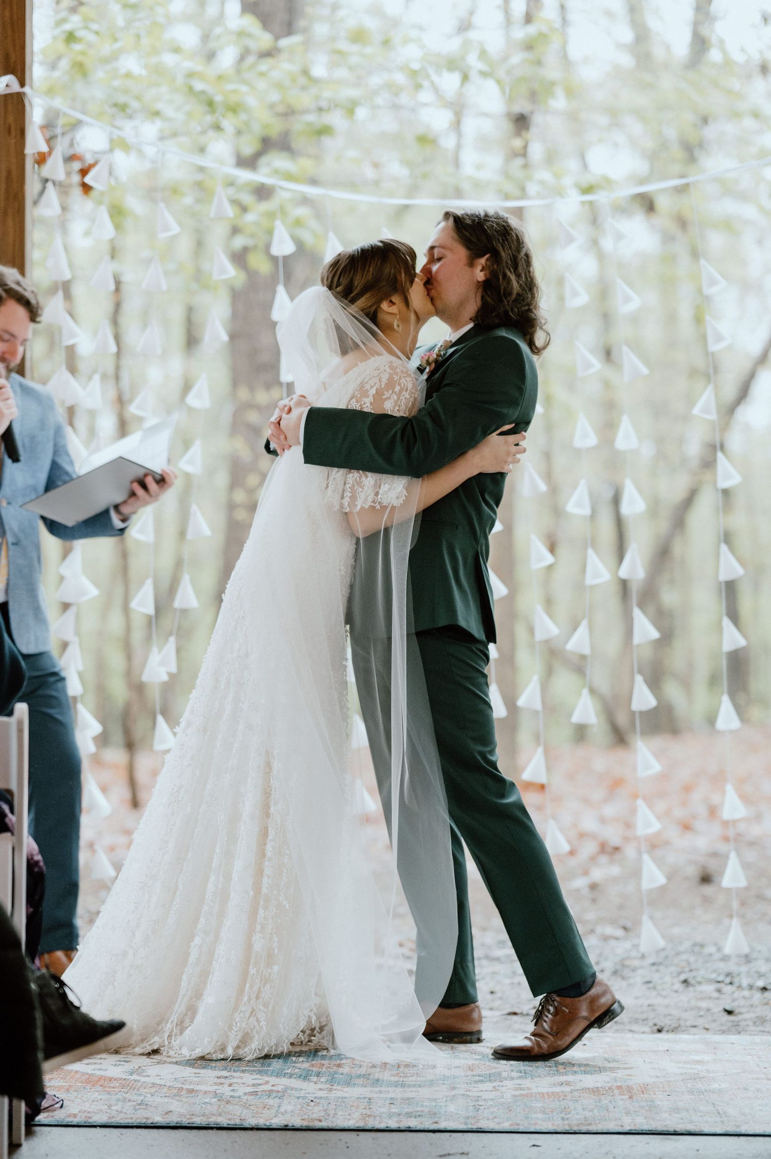 Newlyweds share their first kiss during a woodland wedding ceremony with string lights in the background.
