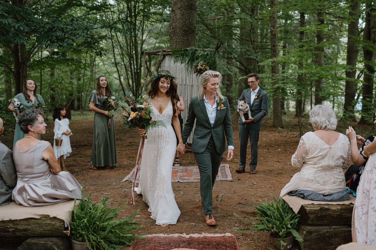 A couple walks down a forest aisle at their outdoor wedding ceremony surrounded by guests.