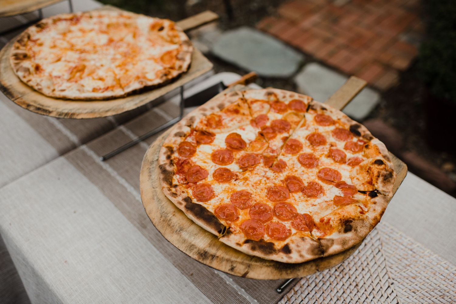 Two freshly baked pepperoni pizzas served on white tablecloth outdoors.