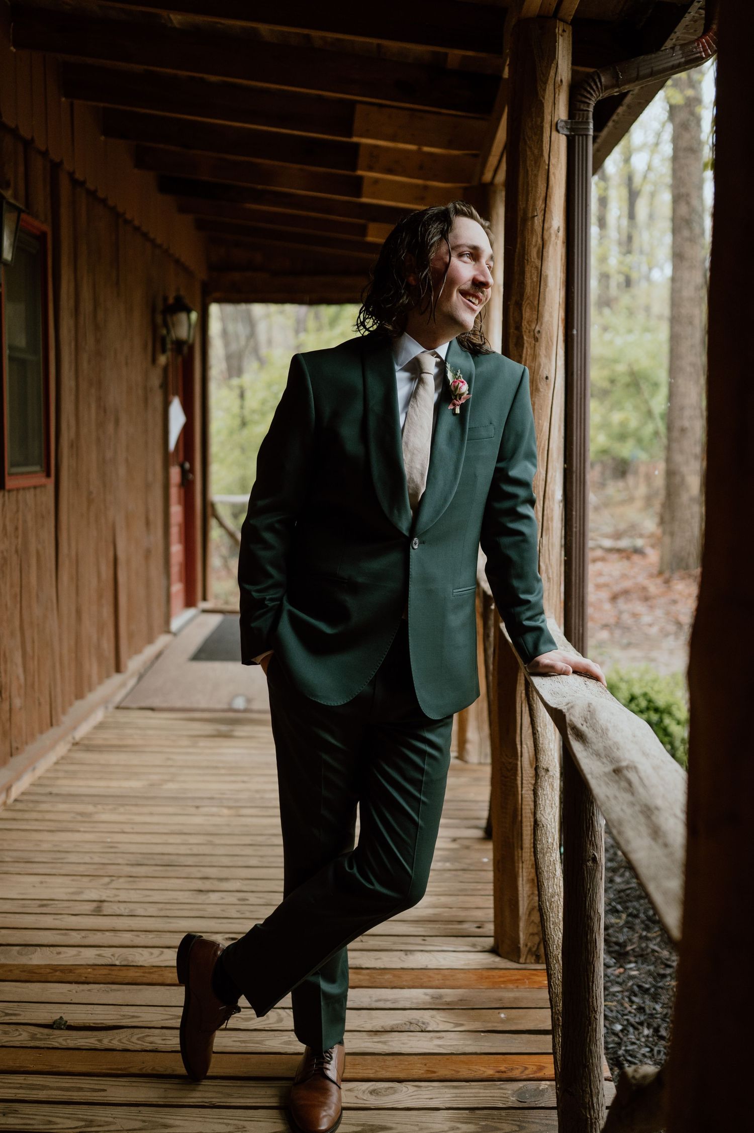A formal portrait of a person in an emerald green suit standing on a rustic wooden porch.