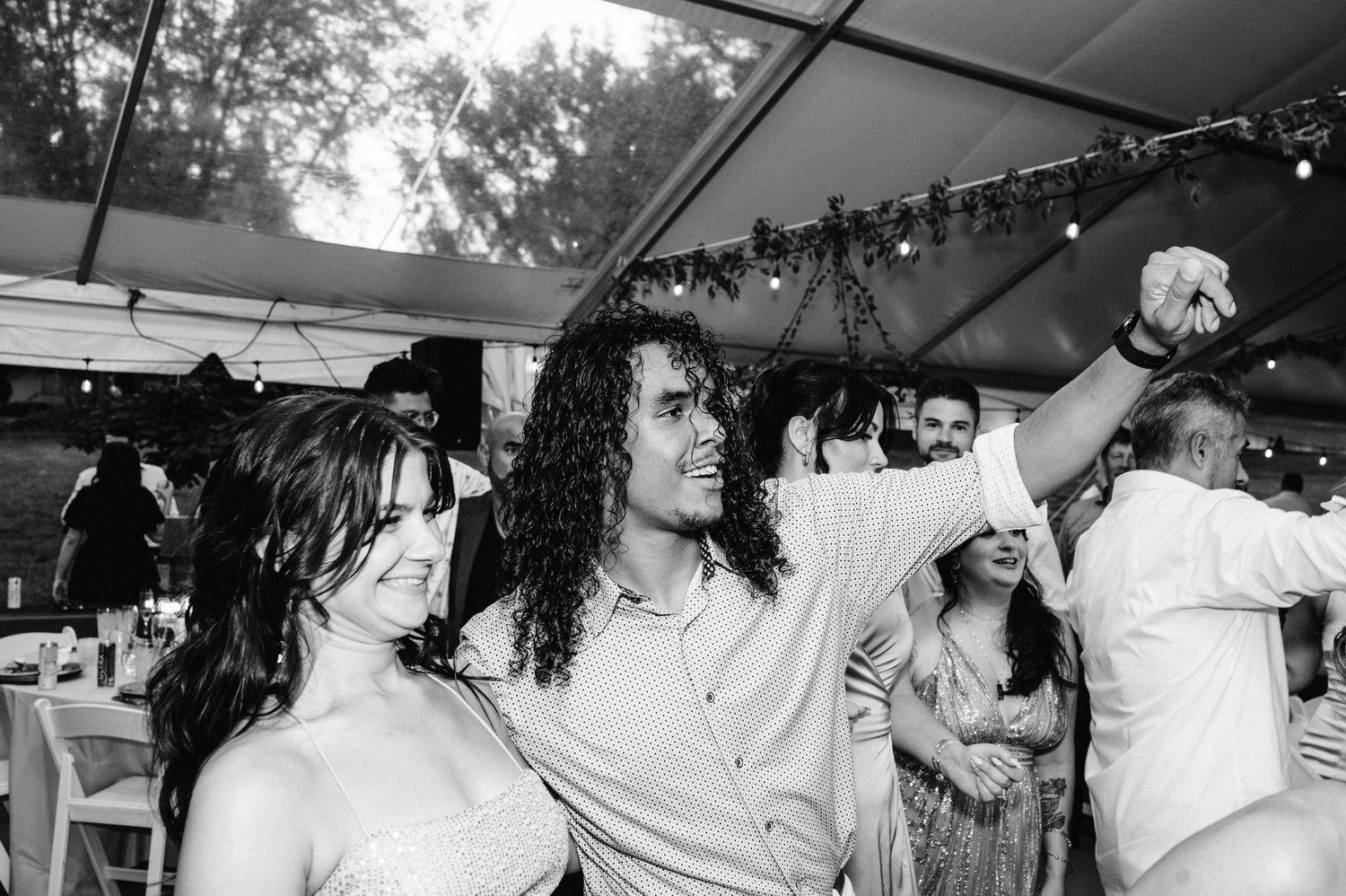 Black and white photo of wedding guests dancing joyfully under a tent.