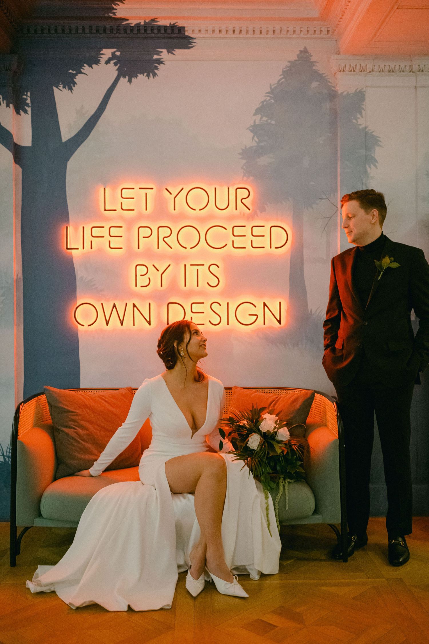 Wedding couple poses beneath neon sign reading 'Let your life proceed by its own design' on orange couch.